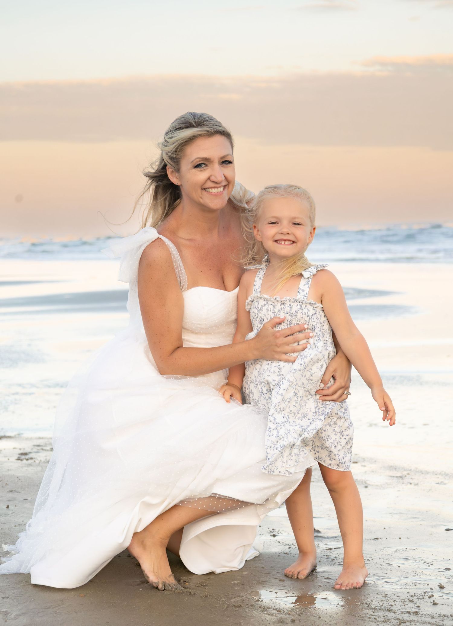 Bride in white gown kneeling beside young girl on sandy beach at sunset with ocean waves in background.