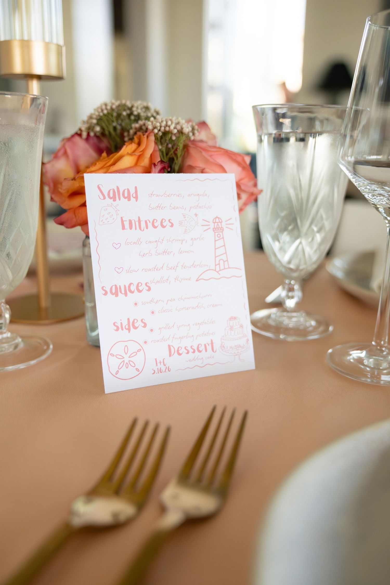 Elegant wedding table setting with coral menu card, gold forks, crystal glasses, and pink floral centerpiece.