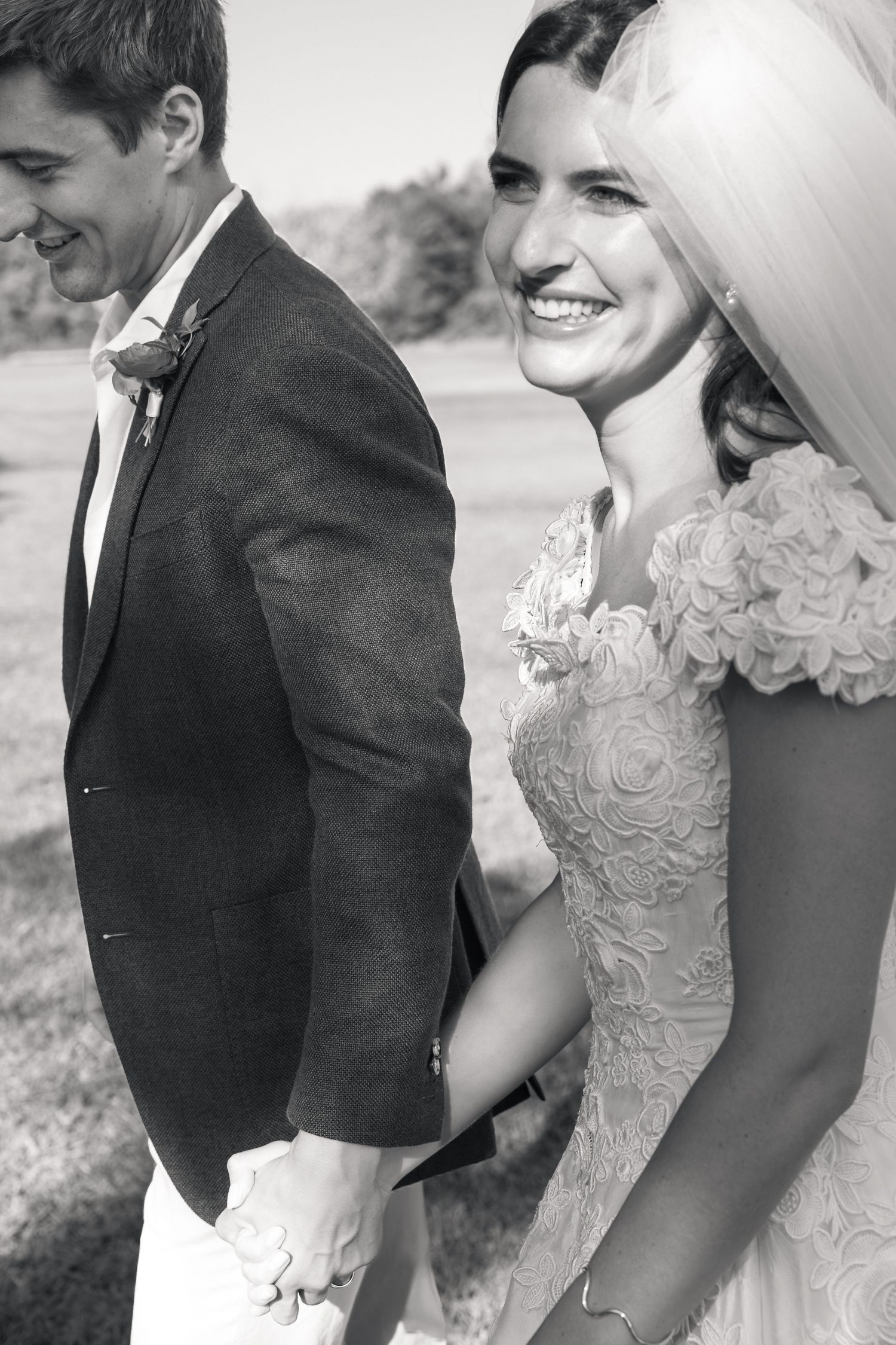 Black and white photo of smiling bride in lace dress holding hands with groom in suit at outdoor wedding.