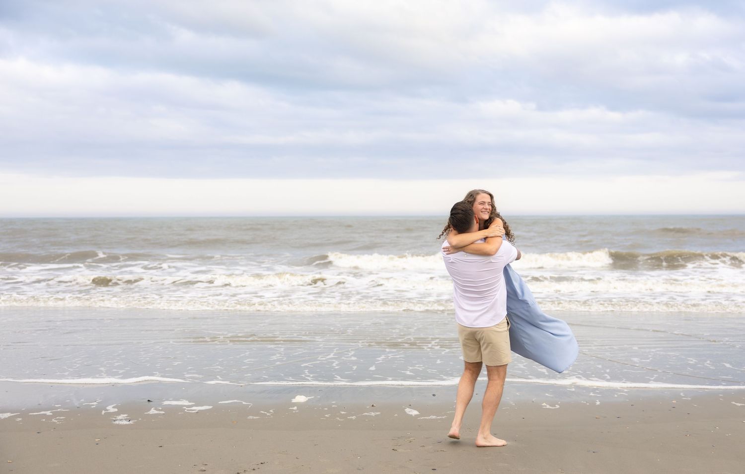 Couple embracing on a sandy beach with ocean waves and cloudy sky in the background.
