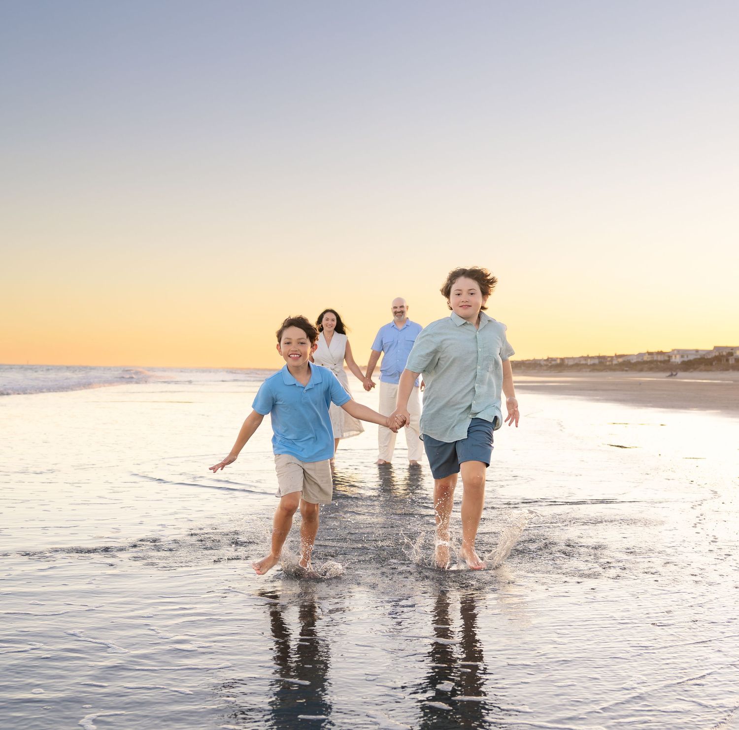 Family of four enjoying a sunset beach walk, two boys running through ocean waves while parents hold hands behind them.