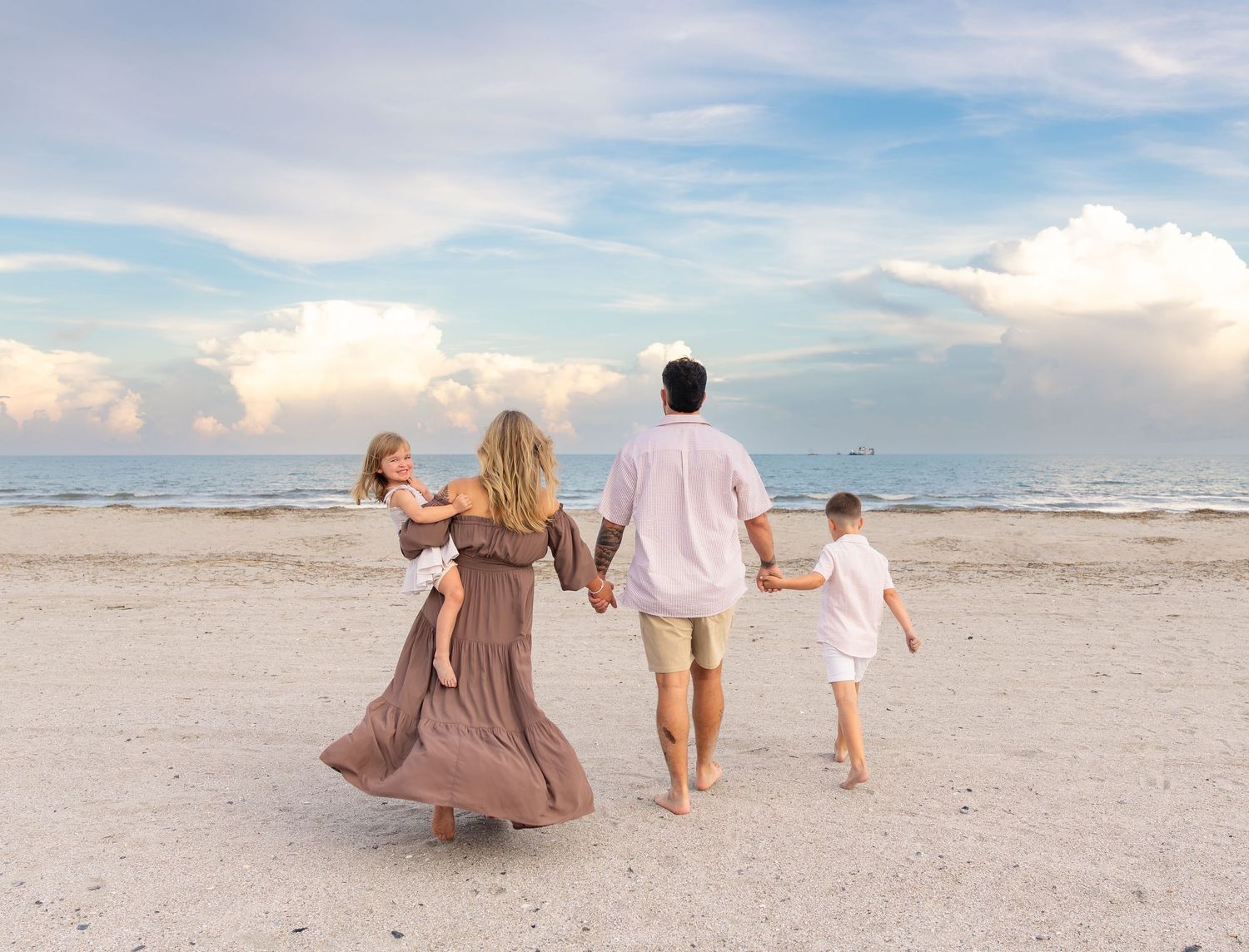 Family of four walking hand-in-hand on a sandy beach at sunset, with ocean waves and dramatic clouds in the background.