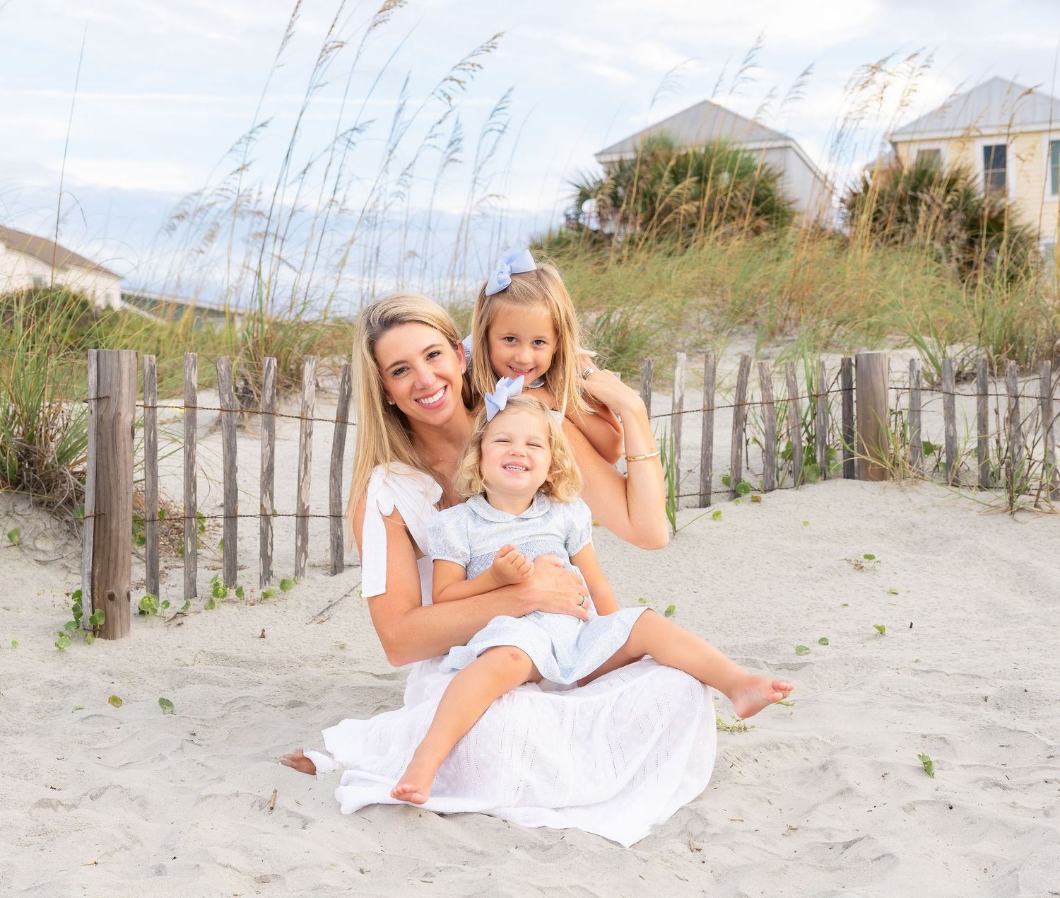 A woman and two young girls smile together on a sandy beach with sea oats and a wooden fence backdrop.
