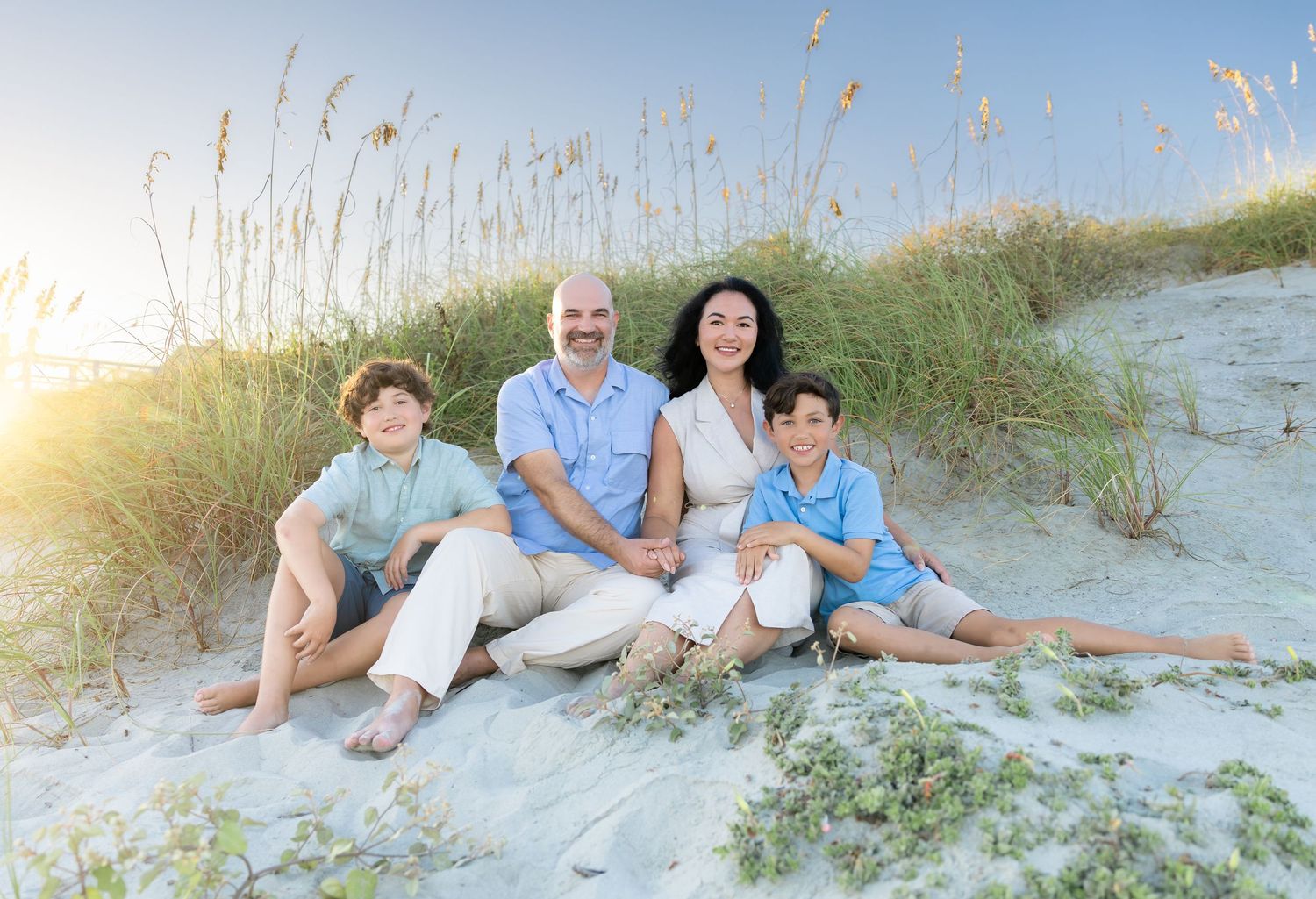 Family of four smiling on sandy beach dunes with sea oats at golden hour.