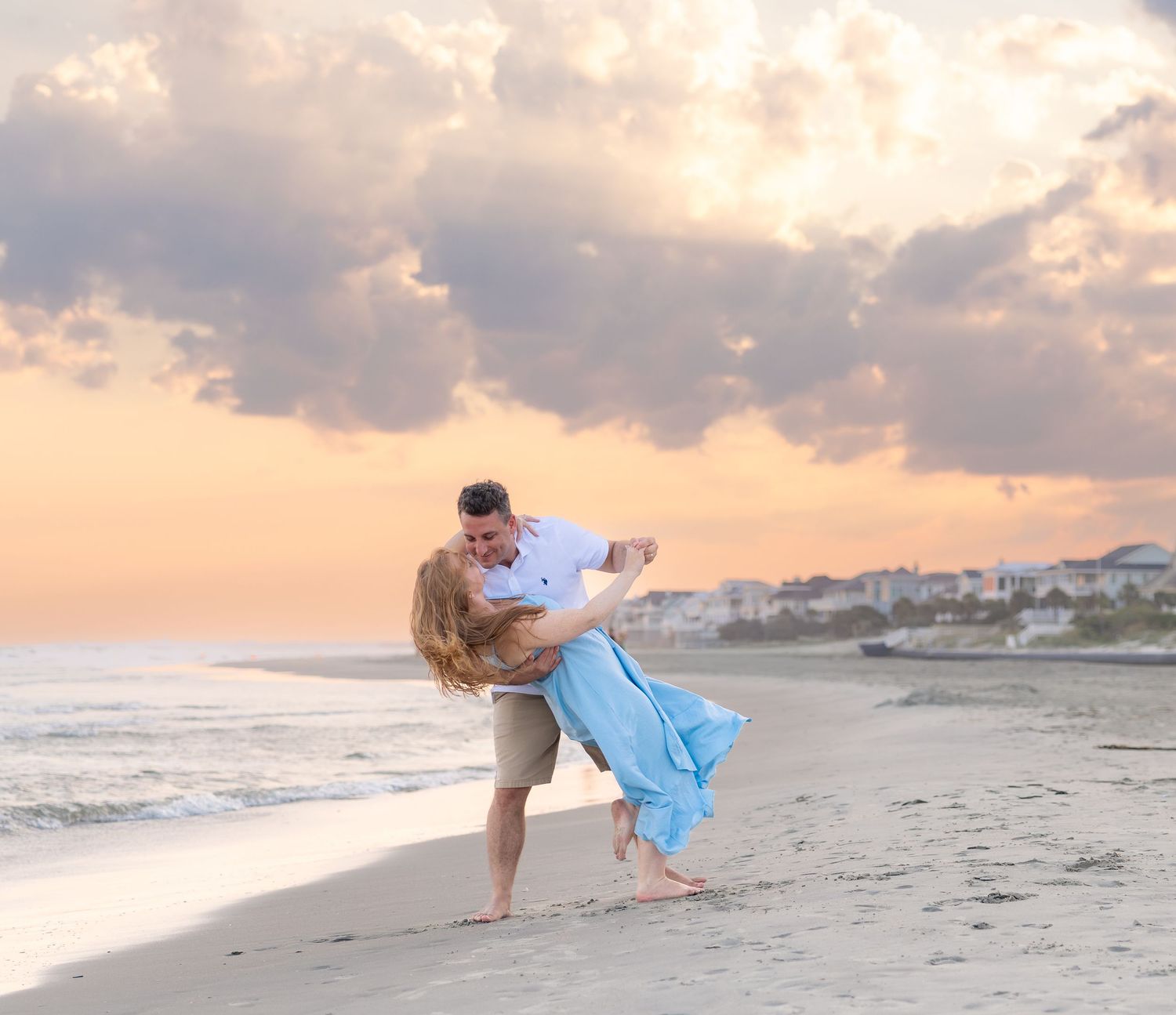 Couple dancing romantically on a sandy beach at sunset with pastel cloudy sky and ocean waves in background.