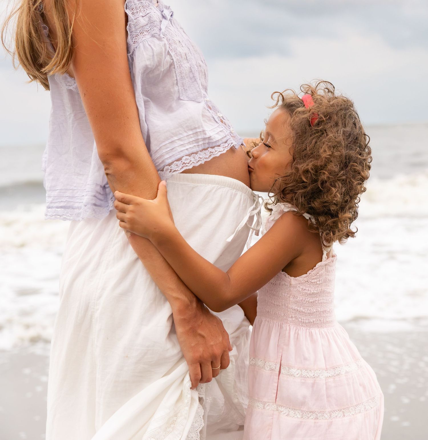 A young girl in a pink dress hugging her pregnant mother on a beach.