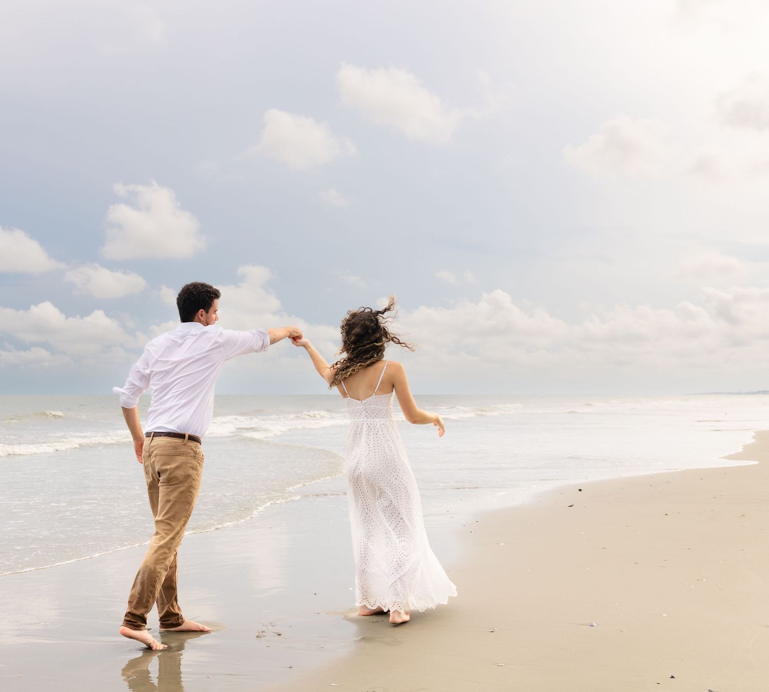 Couple dancing on a sandy beach shore, woman in white dress and man in white shirt holding hands by the ocean waves.