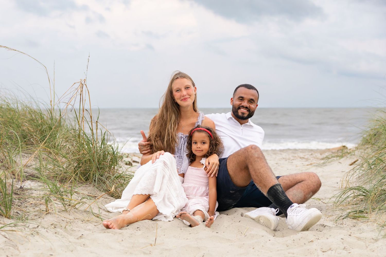 Family of three sitting on sandy beach dunes, smiling couple with young daughter, ocean waves in background.