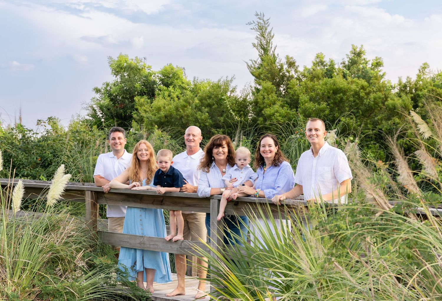 Large family posing together on a wooden deck surrounded by lush greenery at a coastal location.