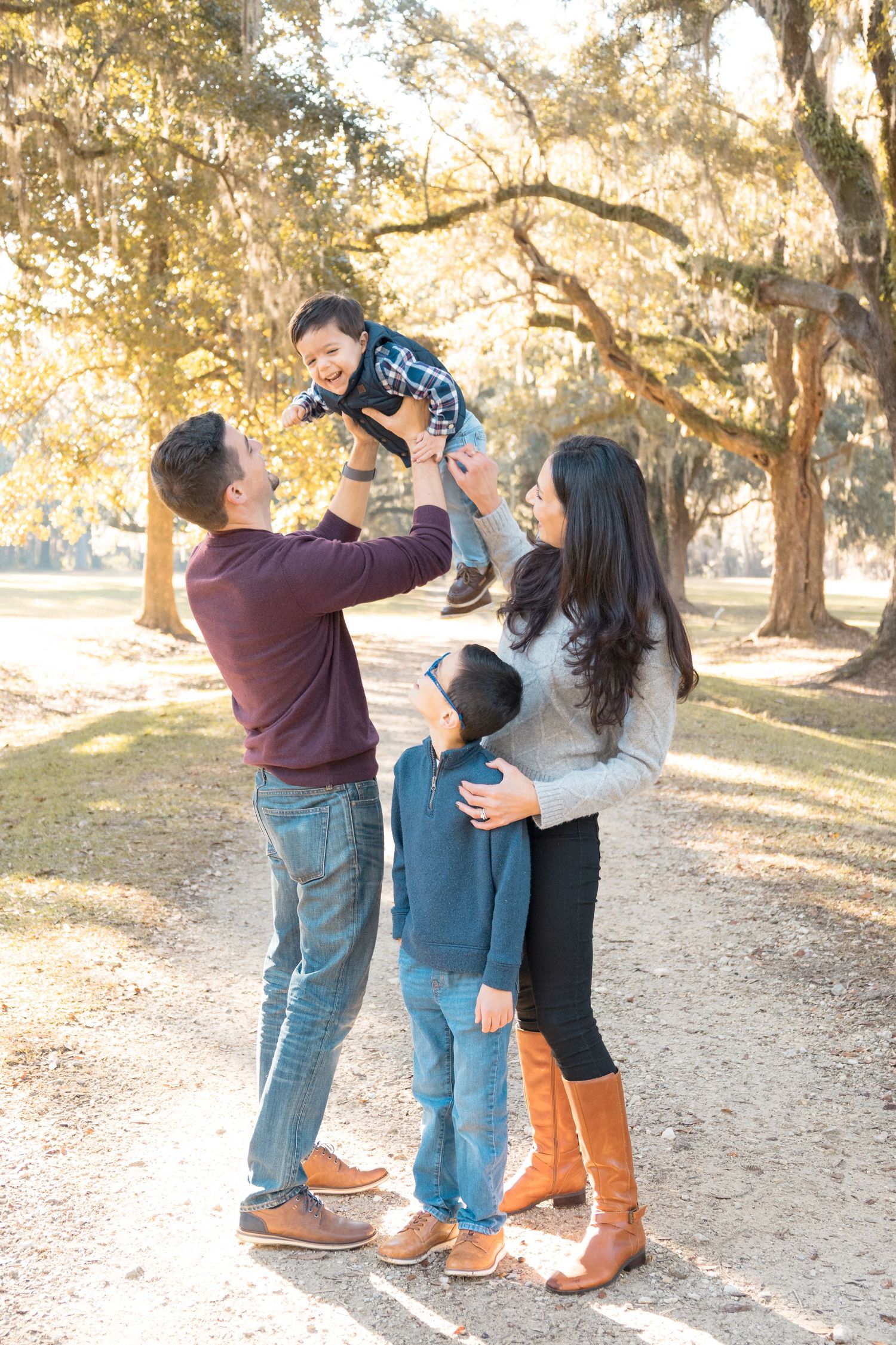 Family of four enjoys an outdoor portrait session as a father lifts a laughing child overhead beneath sunlit oak trees on a gravel path.