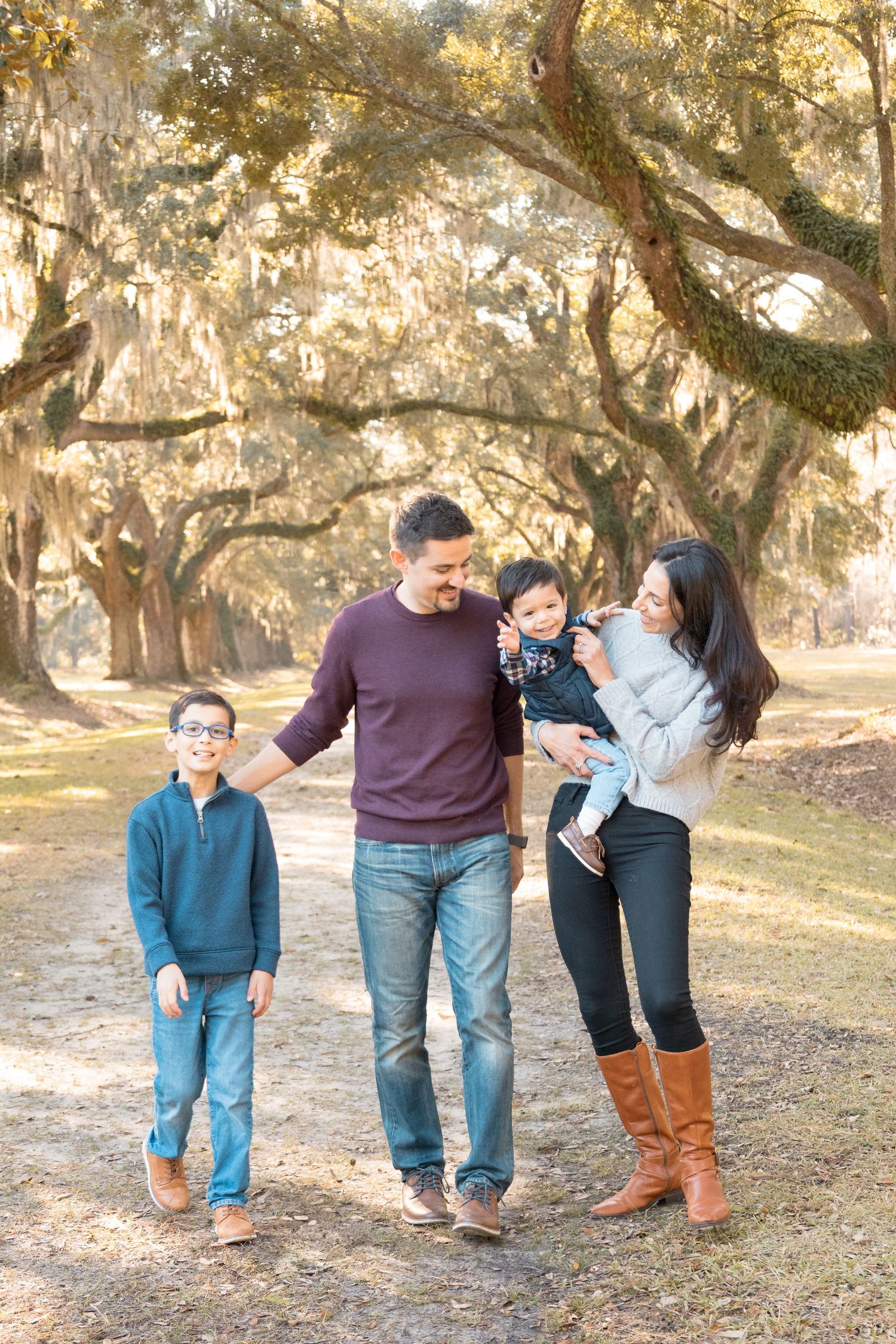 Smiling family of four walking together on a sunlit path beneath live oak trees draped in Spanish moss during an outdoor portrait session.
