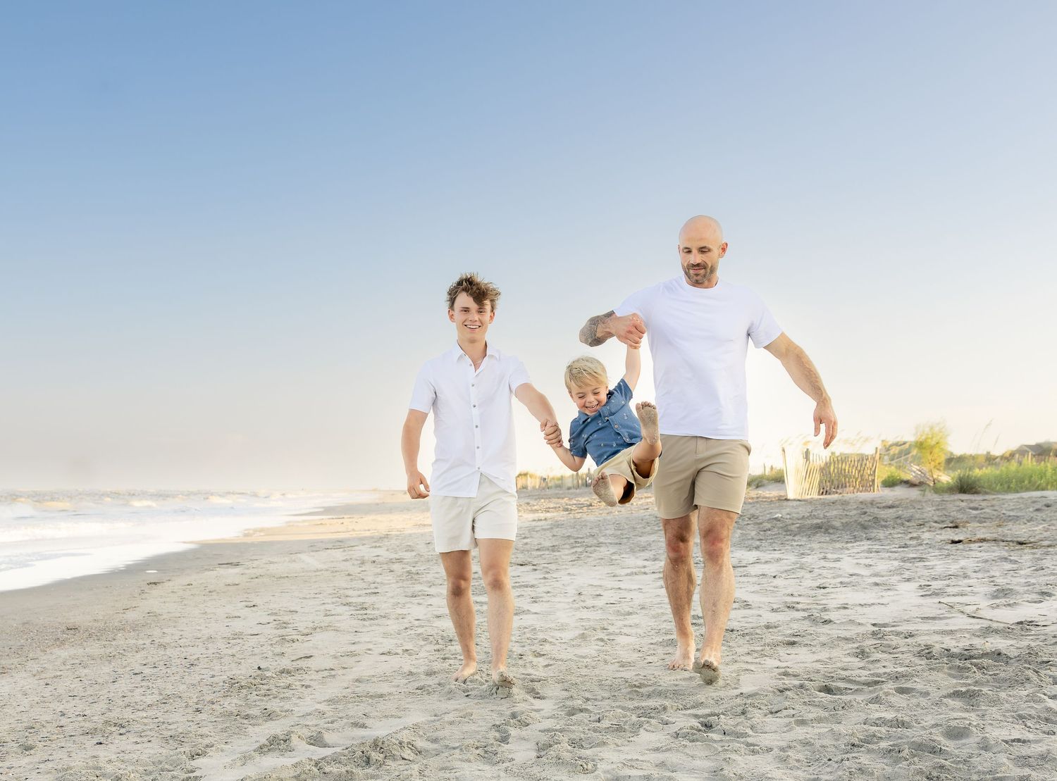 Two dads swing their toddler on a sunny beach, all smiling and barefoot on the sand.