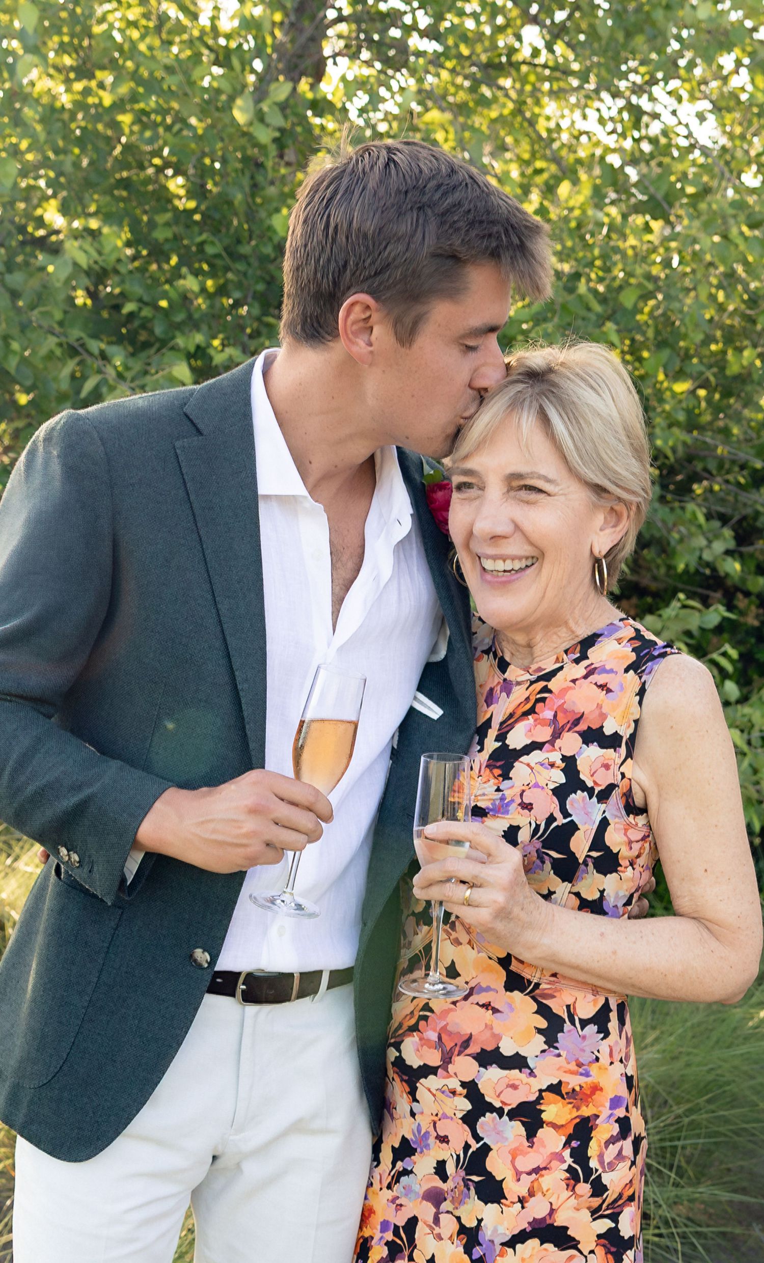 Young man in grey blazer kisses older woman in floral dress while both hold champagne glasses outdoors.