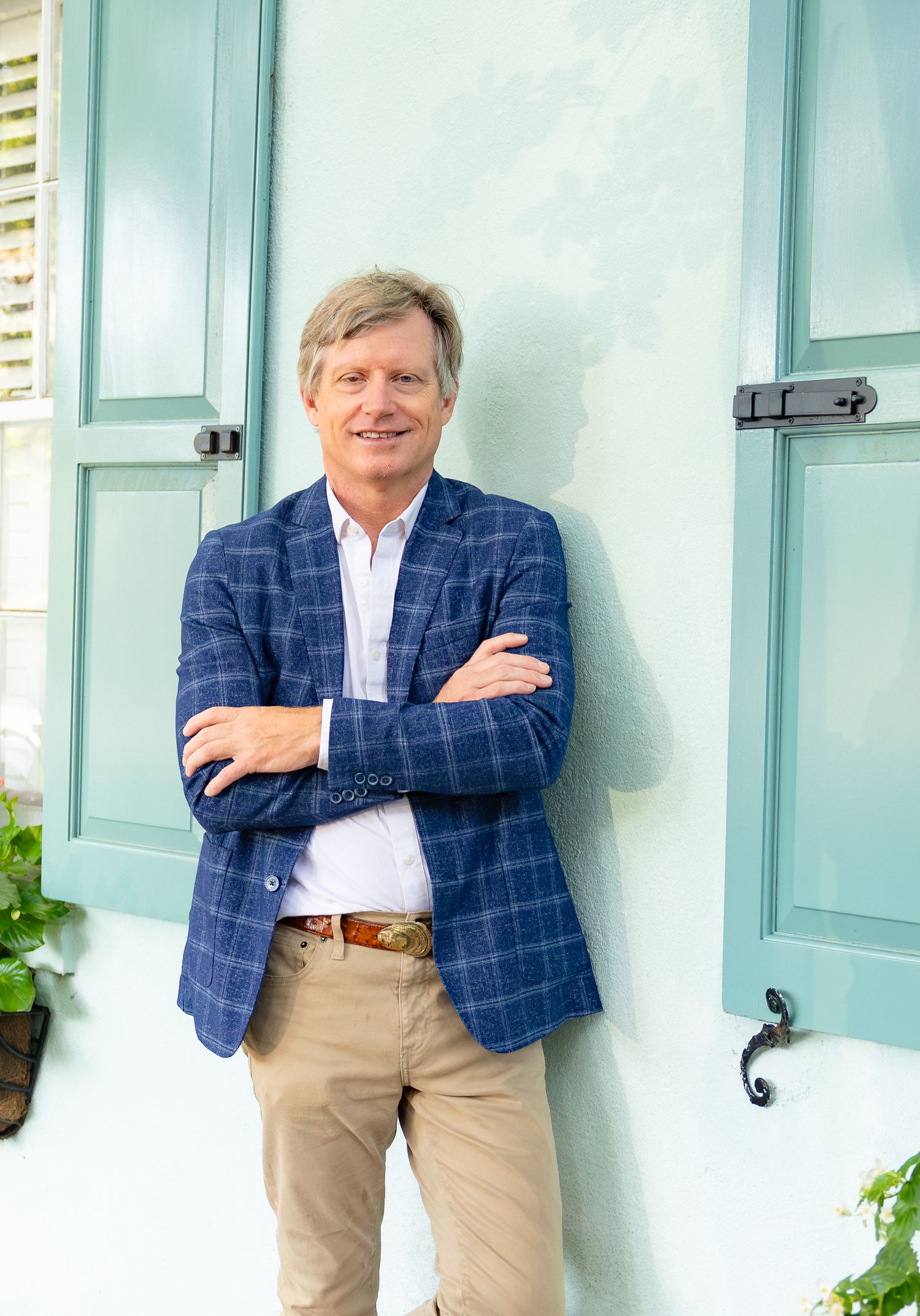 Smiling man in blue plaid blazer and khakis leaning against a turquoise wall with shutters, arms crossed.