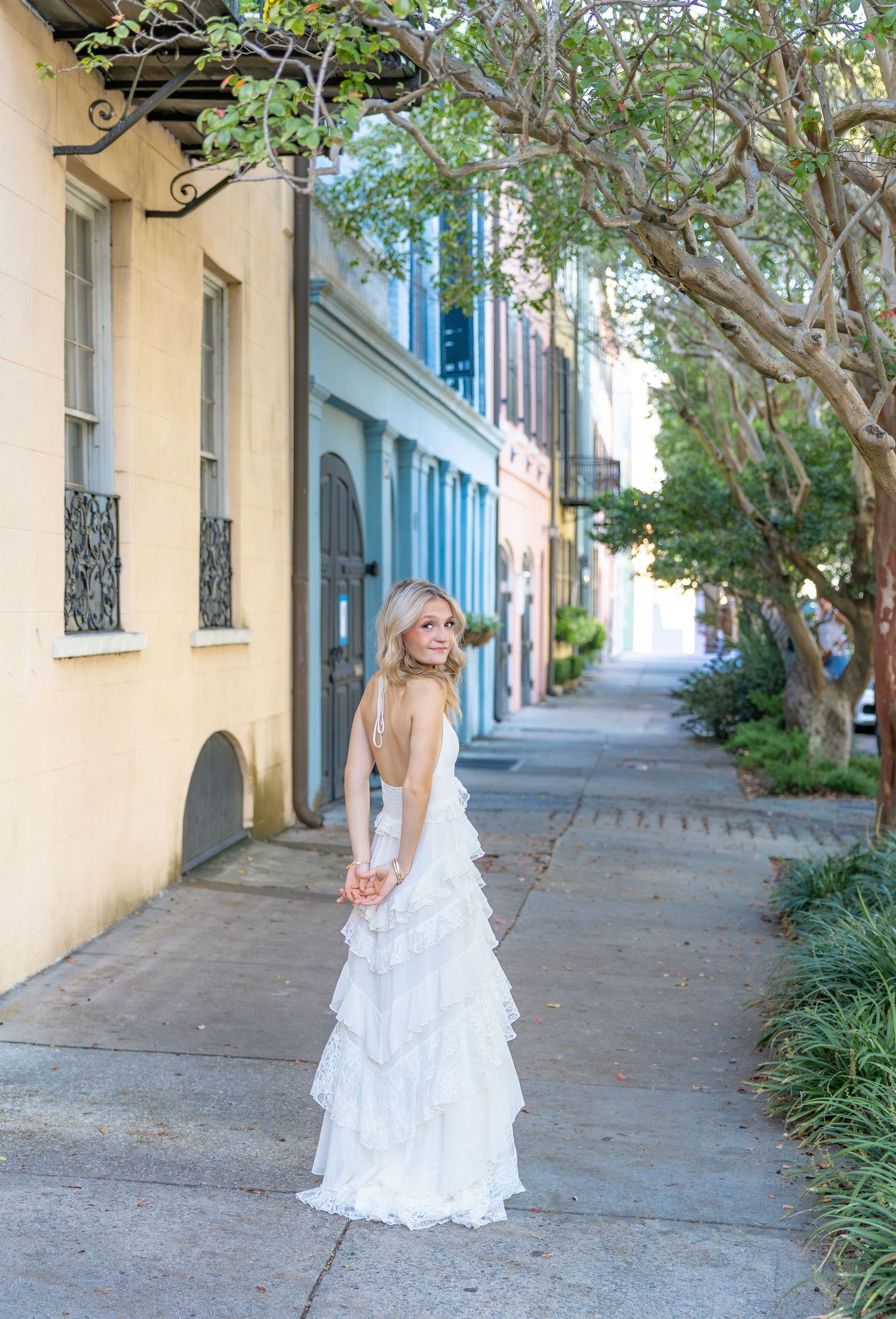 Woman in a white tiered ruffle gown looks over her shoulder on a tree-lined sidewalk beside pastel-colored historic buildings.
