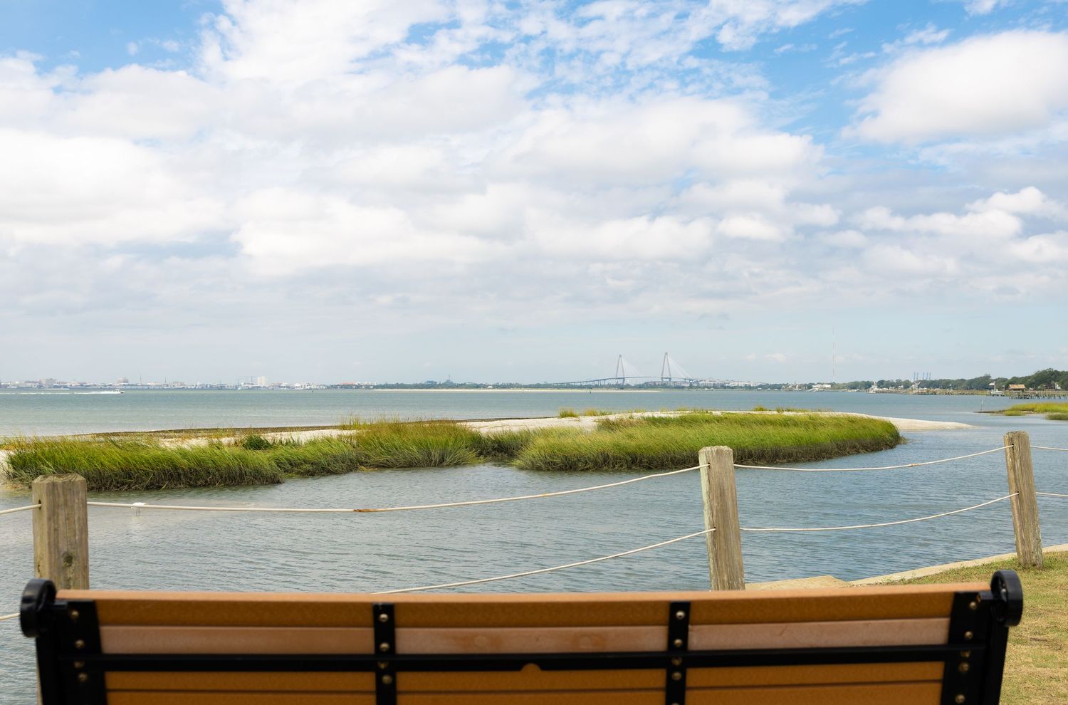 A wooden bench overlooks a serene coastal marsh with grassy islands, calm water, and a partly cloudy sky.