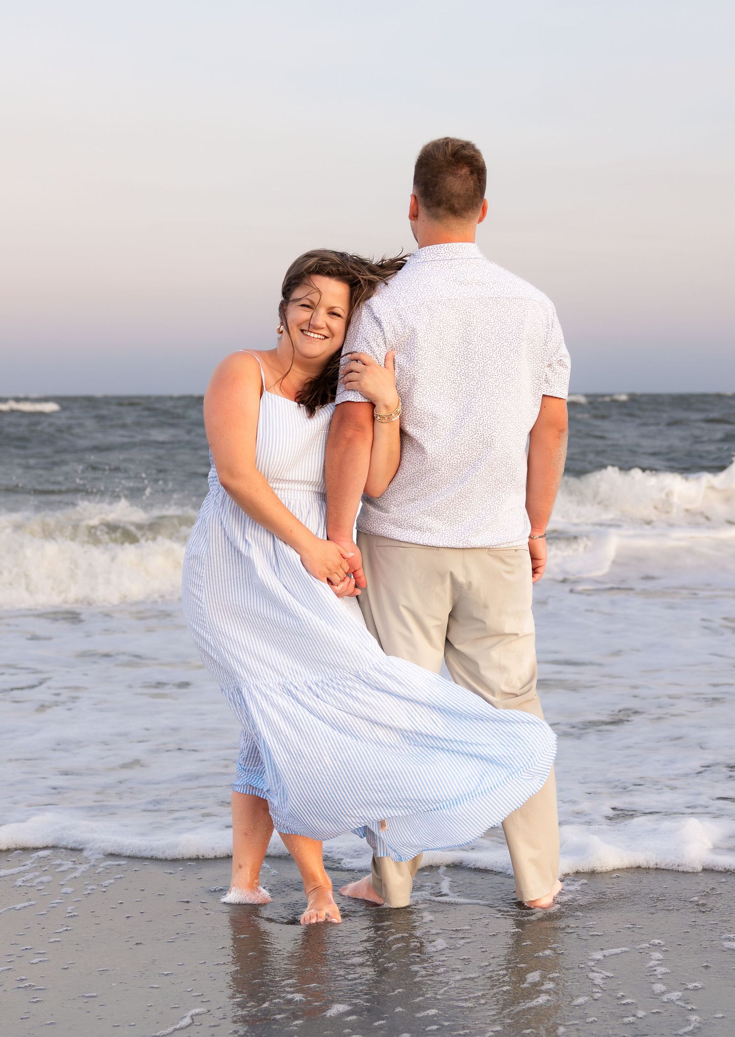 Smiling woman in a flowing white dress leans against a man in a white shirt as they stand in ocean waves on the beach at sunset.