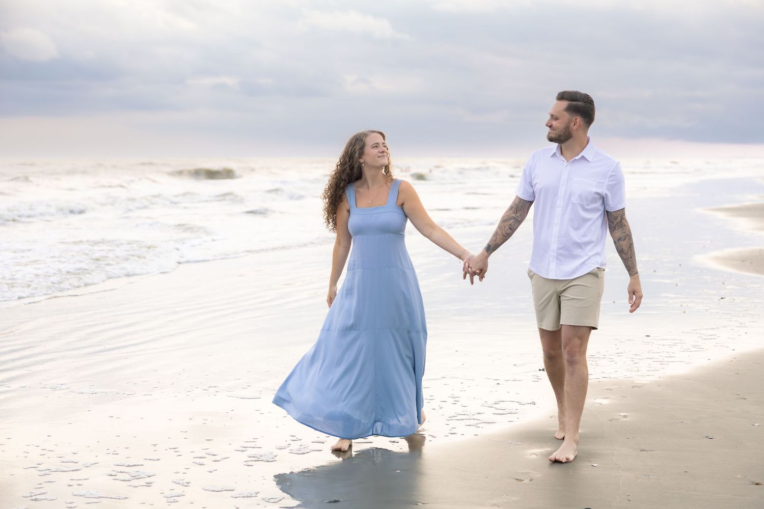 A couple walks hand-in-hand along a sandy beach, the woman in a flowing blue dress, the man in casual shorts and white shirt.