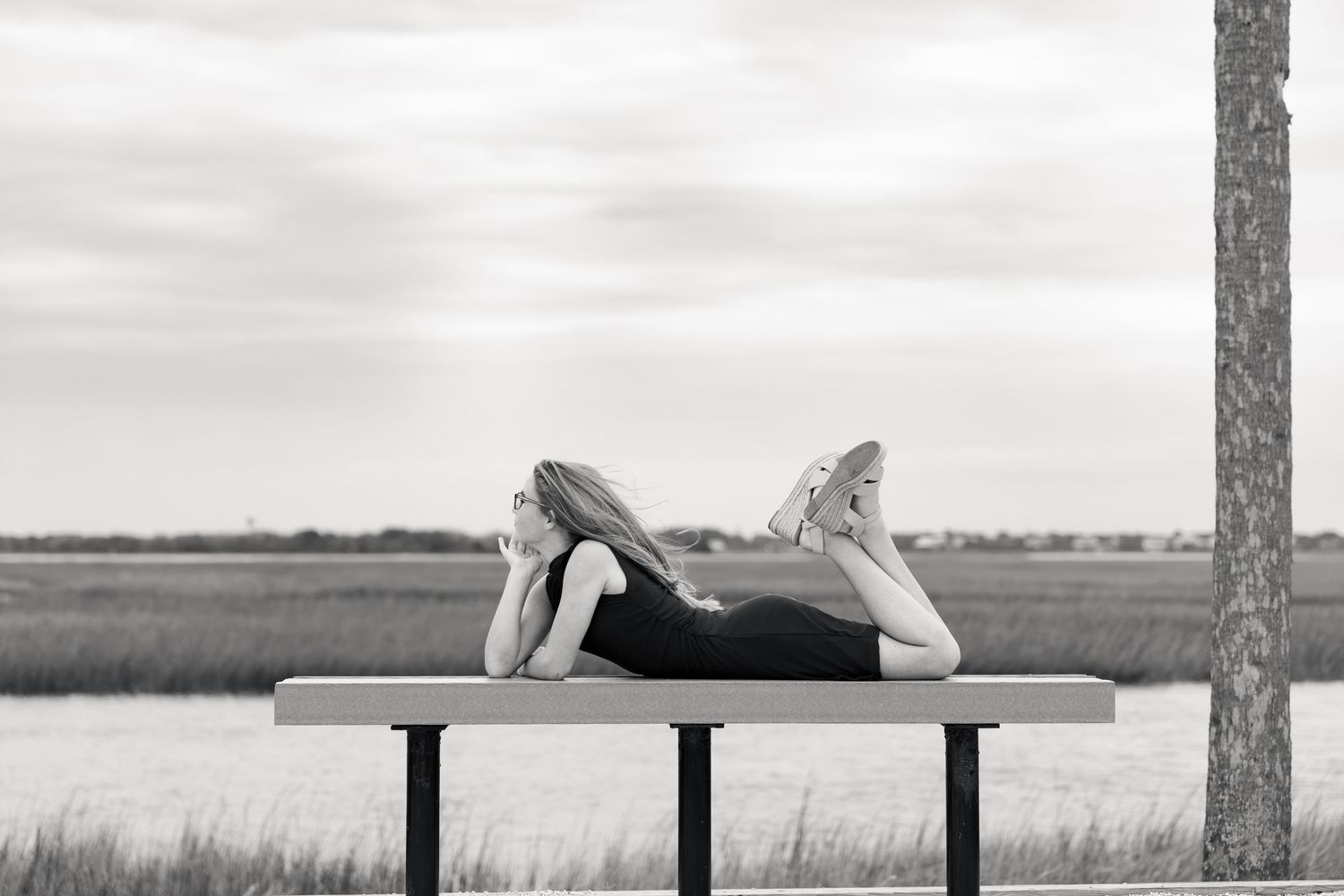 Black and white photo of a woman lying on her stomach on a wooden bench beside a marsh with water, grass and a tall tree.