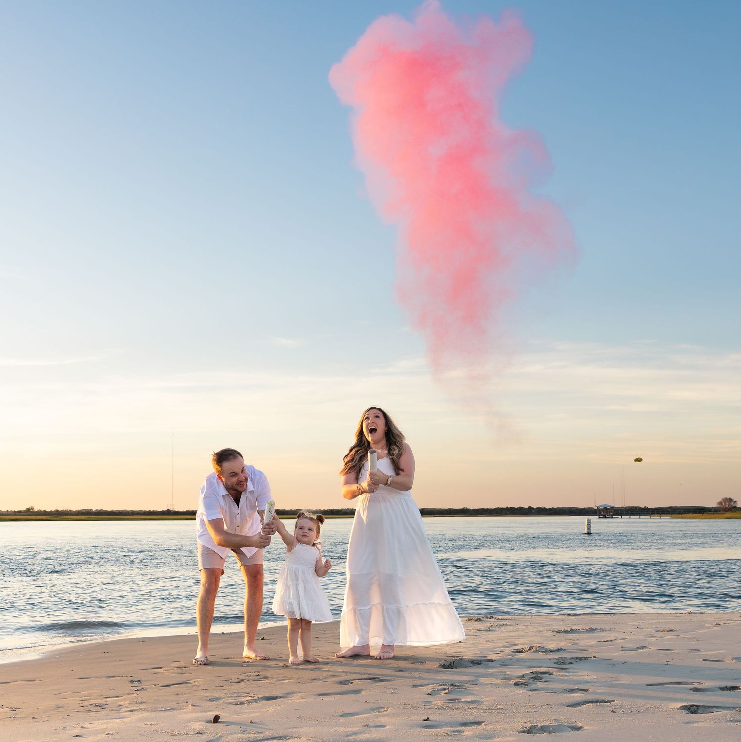 Family gender reveal on beach with pink smoke bomb at sunset, parents and toddler celebrating a baby girl.