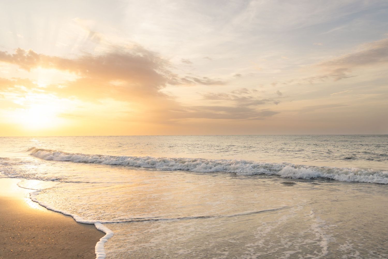 Golden sunrise over calm ocean waves gently washing onto a sandy beach under a soft pastel sky.