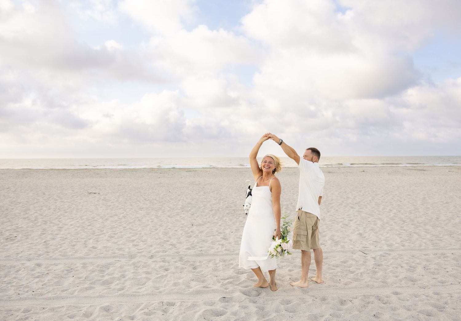 Newlywed couple dancing on a sandy beach in wedding attire, bride holding bouquet under cloudy sky.