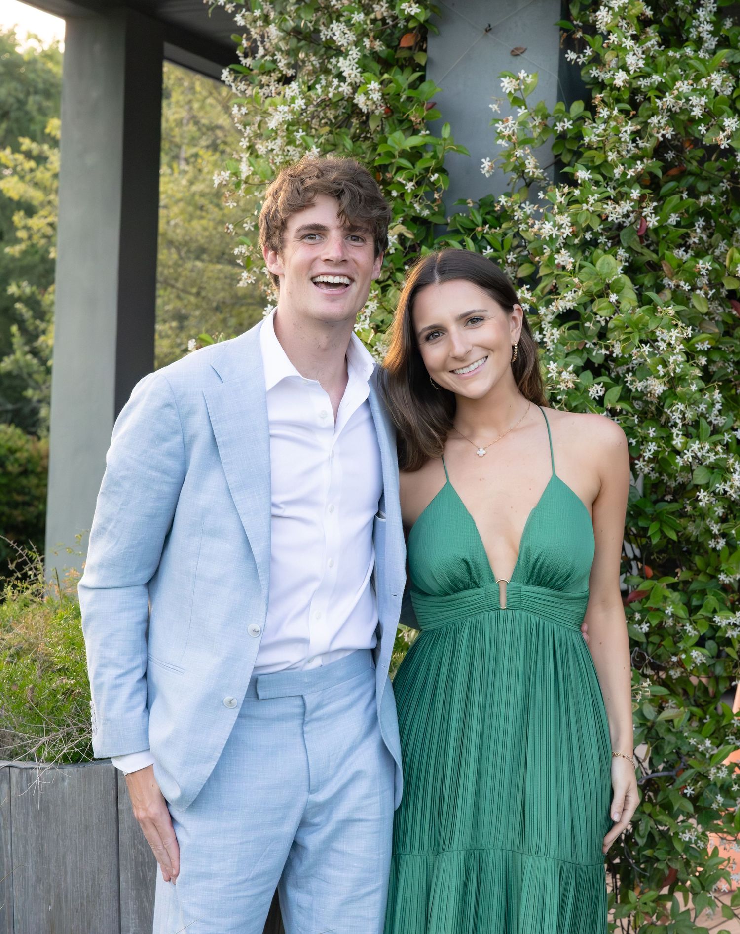 A smiling couple poses outdoors, man in light blue suit and woman in elegant green dress, surrounded by white flowering vines.