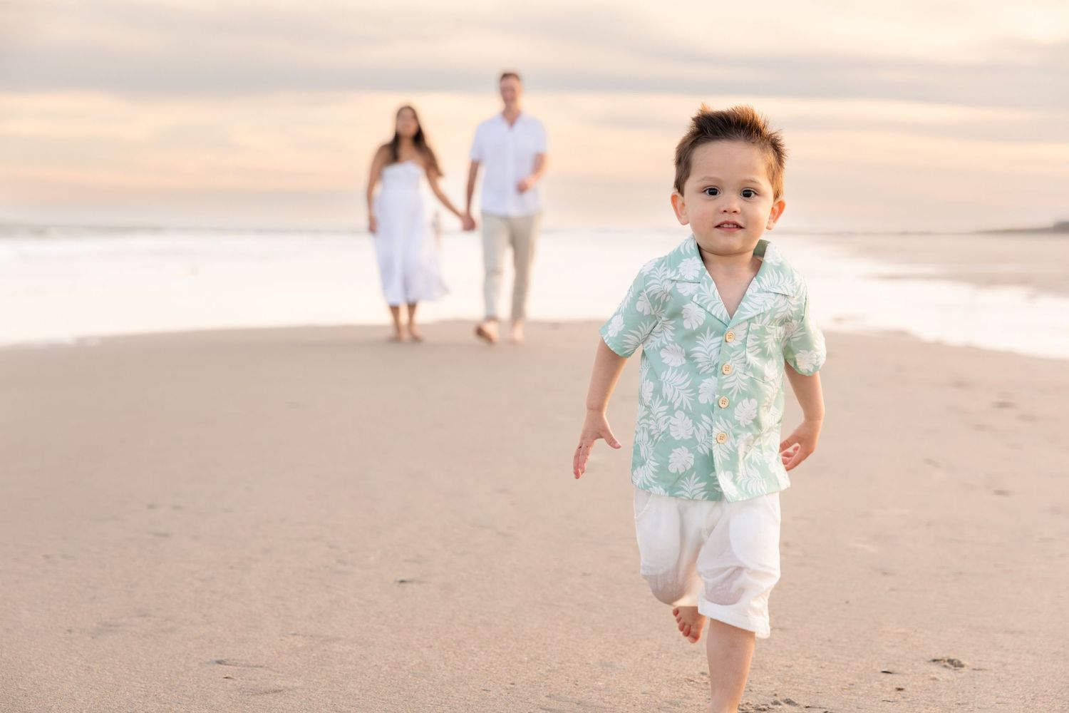 A toddler boy runs on a sandy beach while his parents hold hands in the background at sunset.