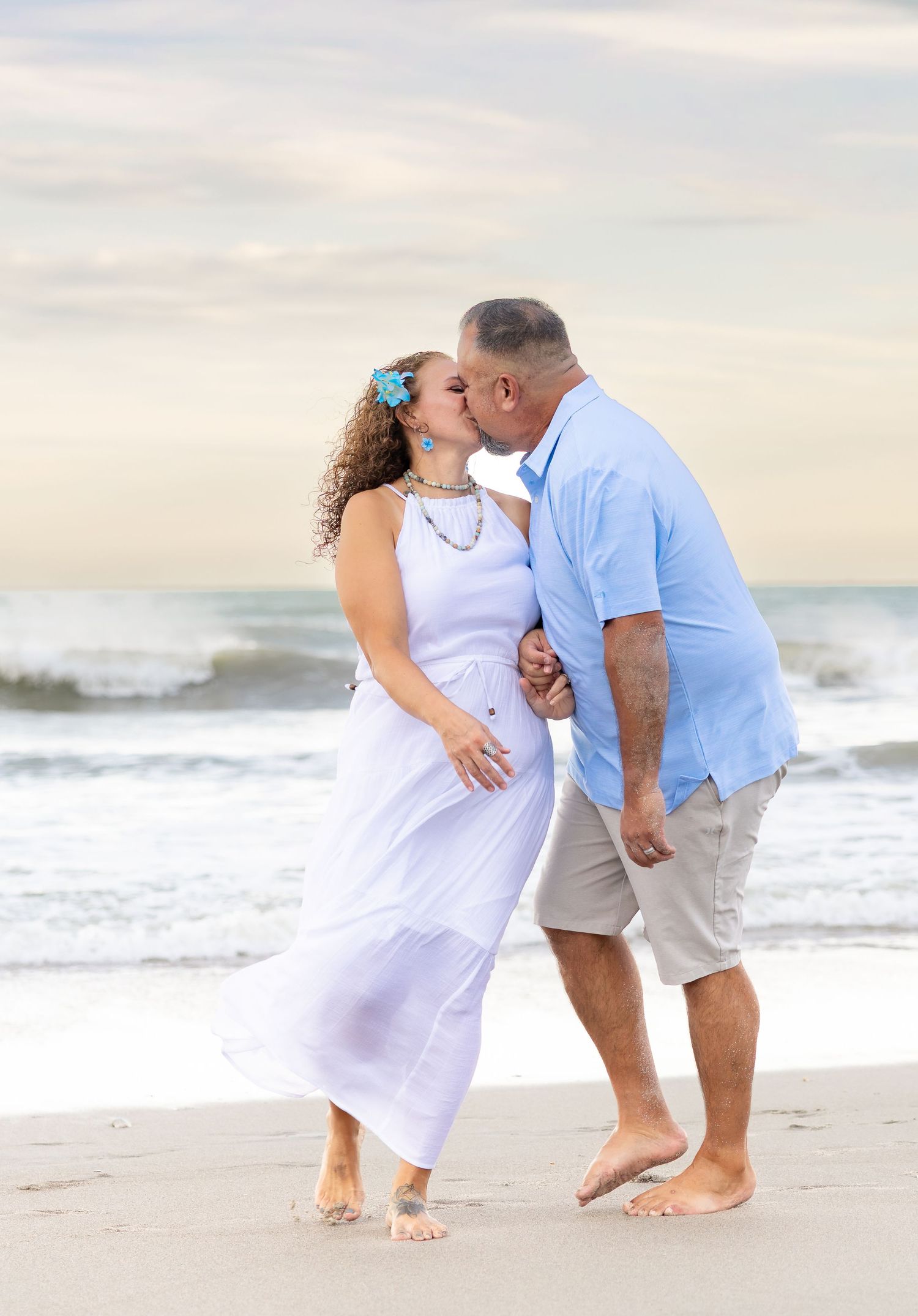 Couple sharing a romantic kiss on a sandy beach, woman in white dress with blue flower, man in light blue shirt.