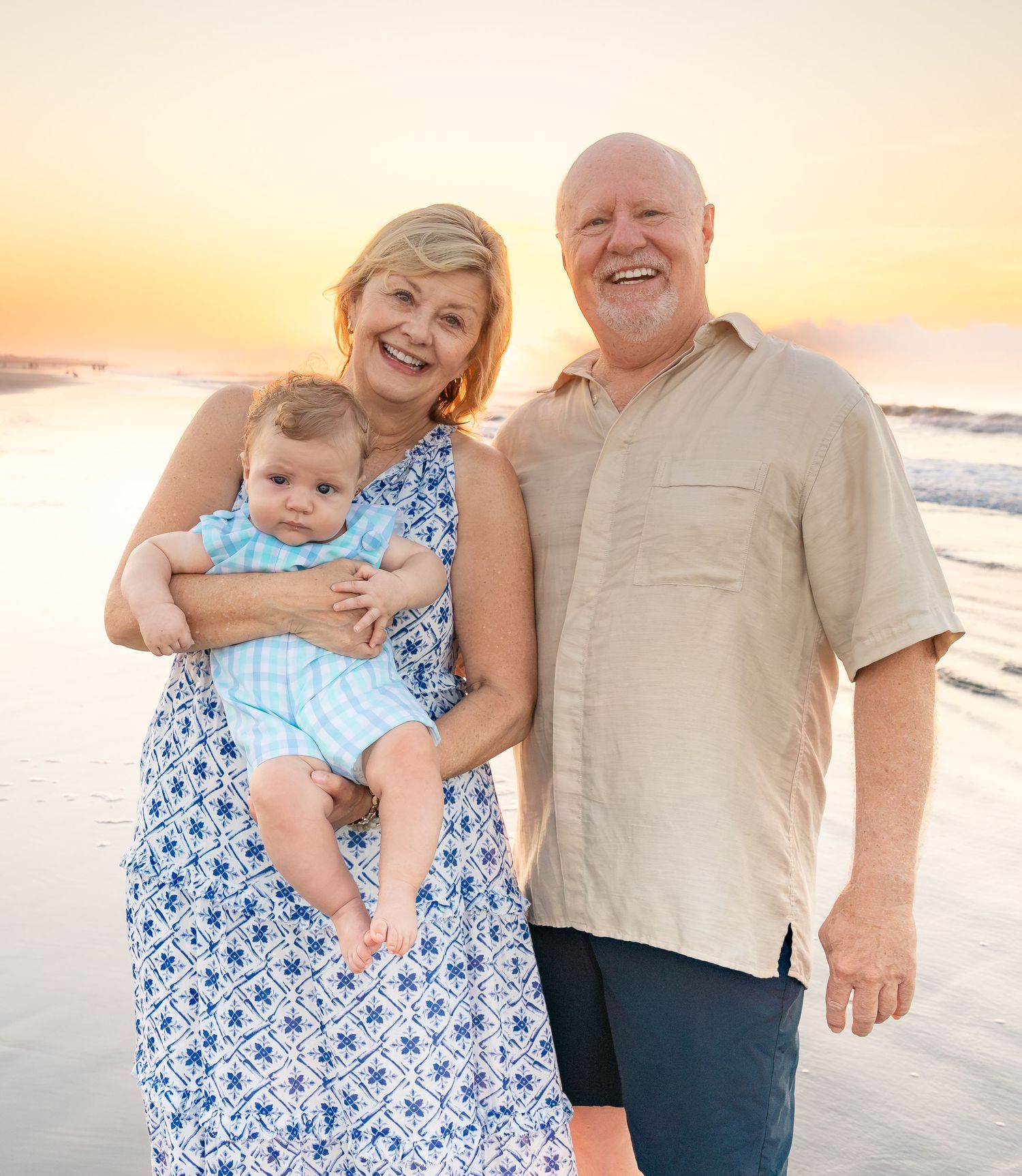 A smiling couple poses with a baby at sunset on the beach, woman in blue dress, man in beige shirt.