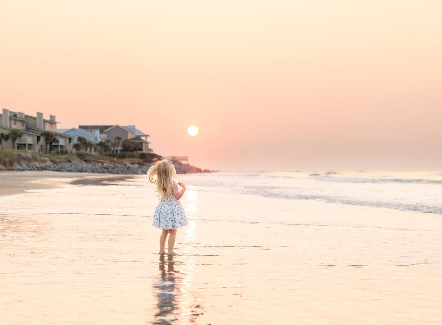 A young girl in a floral dress stands on a sandy beach at sunset, waves gently lapping at her feet.