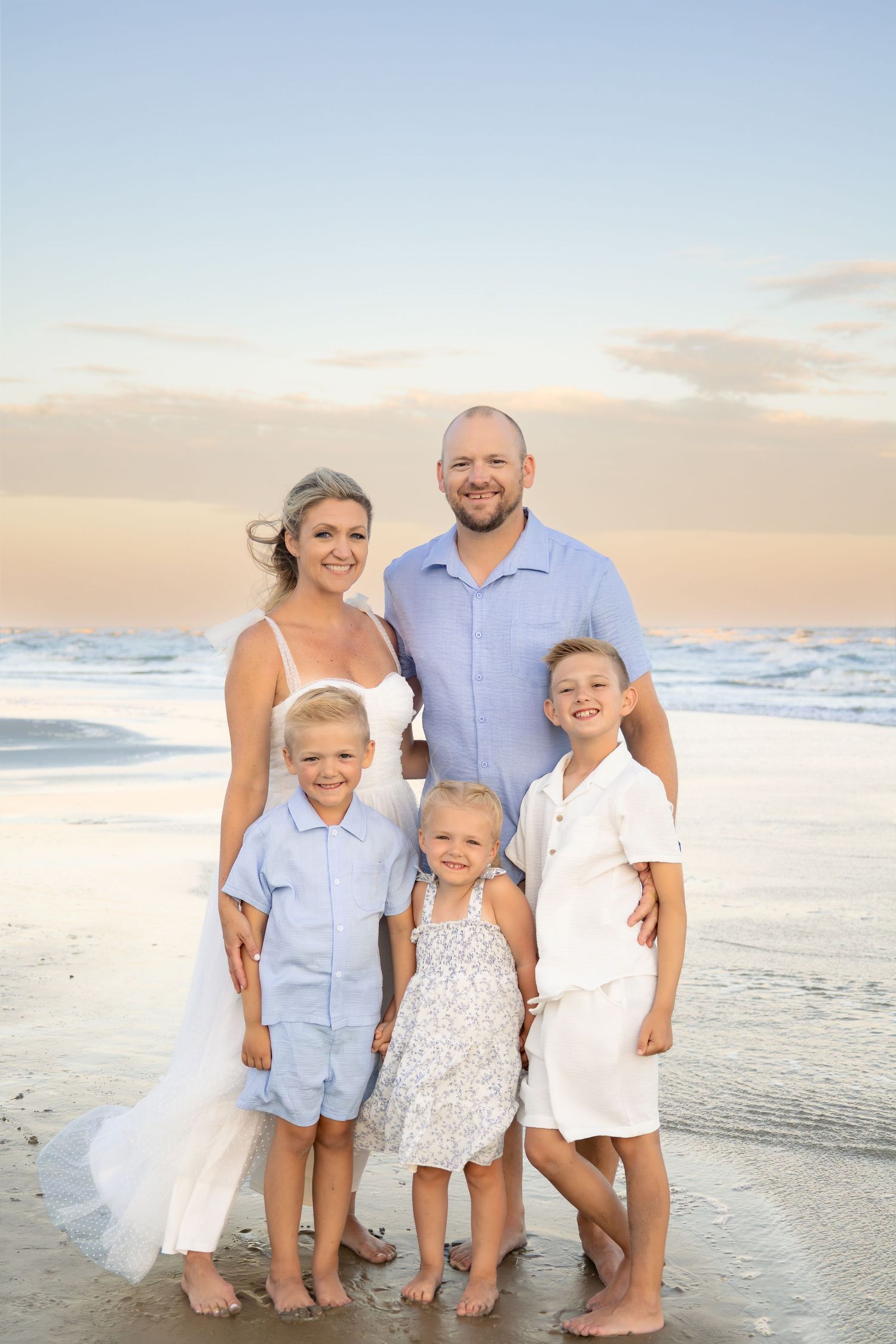 Family of five posing on a beach at sunset, parents and three children dressed in white and blue outfits.