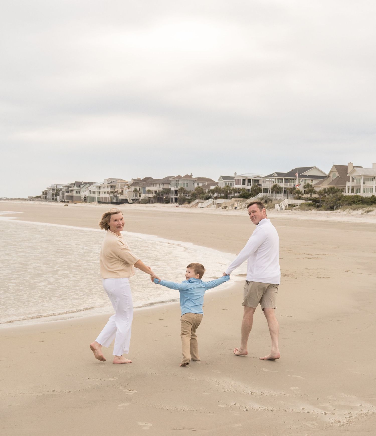 Family of three holding hands and playing on a sandy beach with coastal homes in the background.