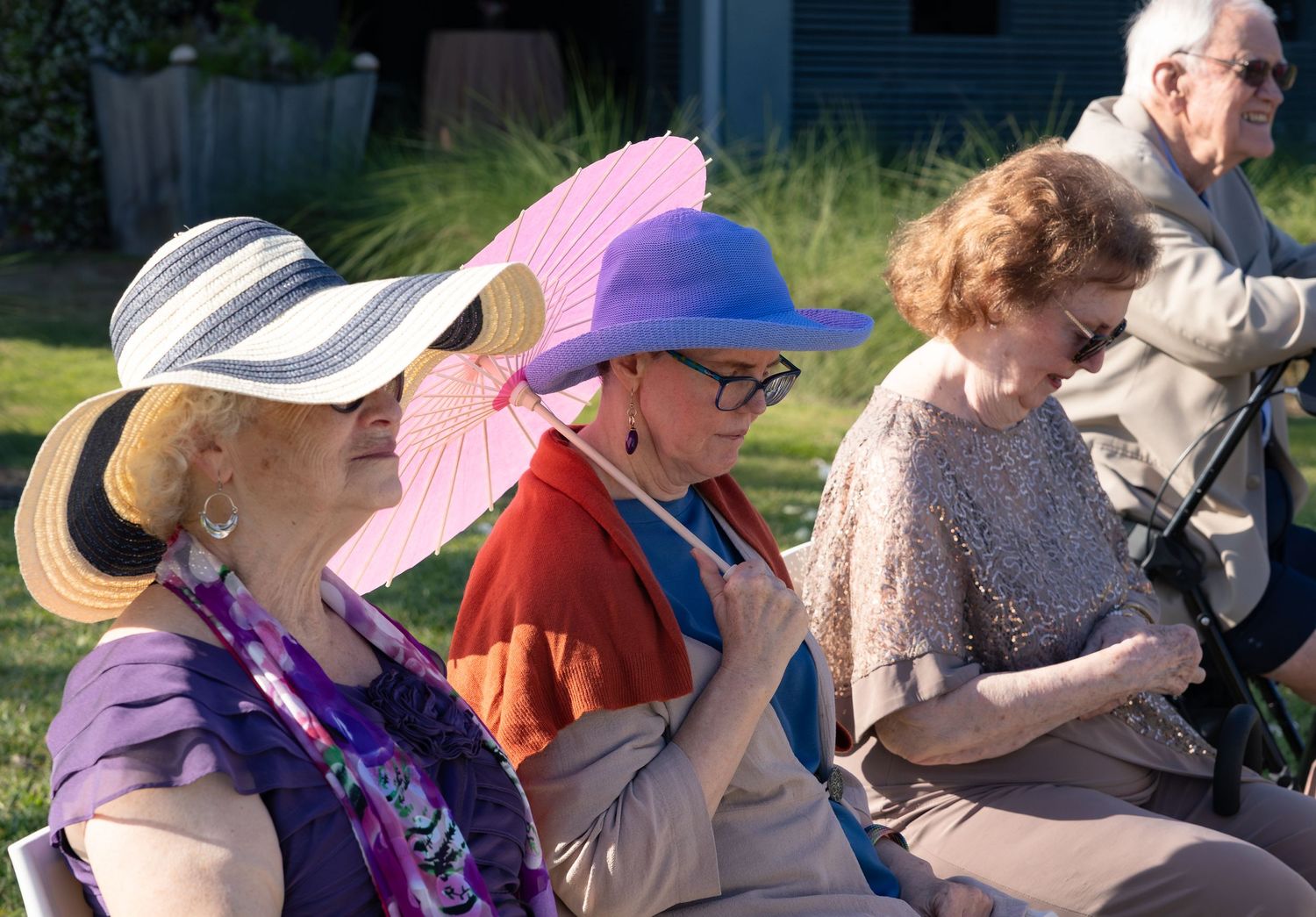 Elderly women wearing colorful sun hats and a pink parasol seated outdoors at a sunny garden event.