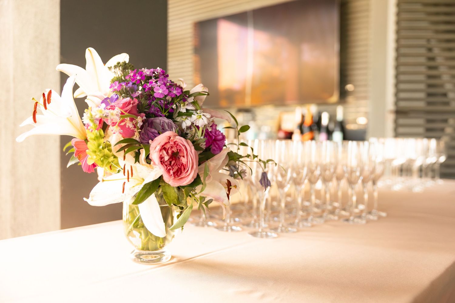 Colorful floral centerpiece with roses and lilies beside rows of champagne glasses on a banquet table.