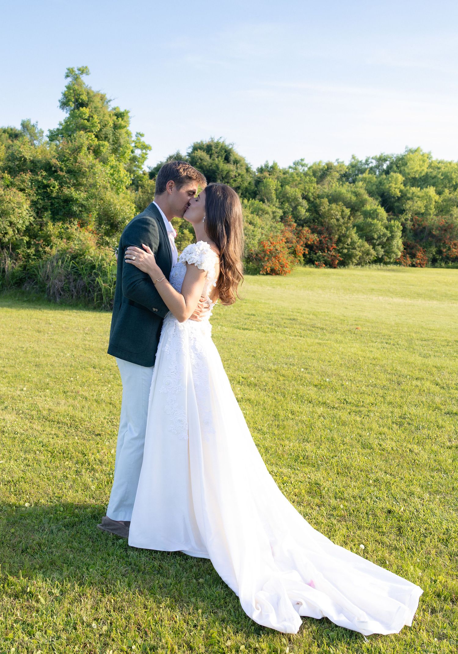 Bride and groom sharing a romantic kiss on a green lawn with trees in the background.