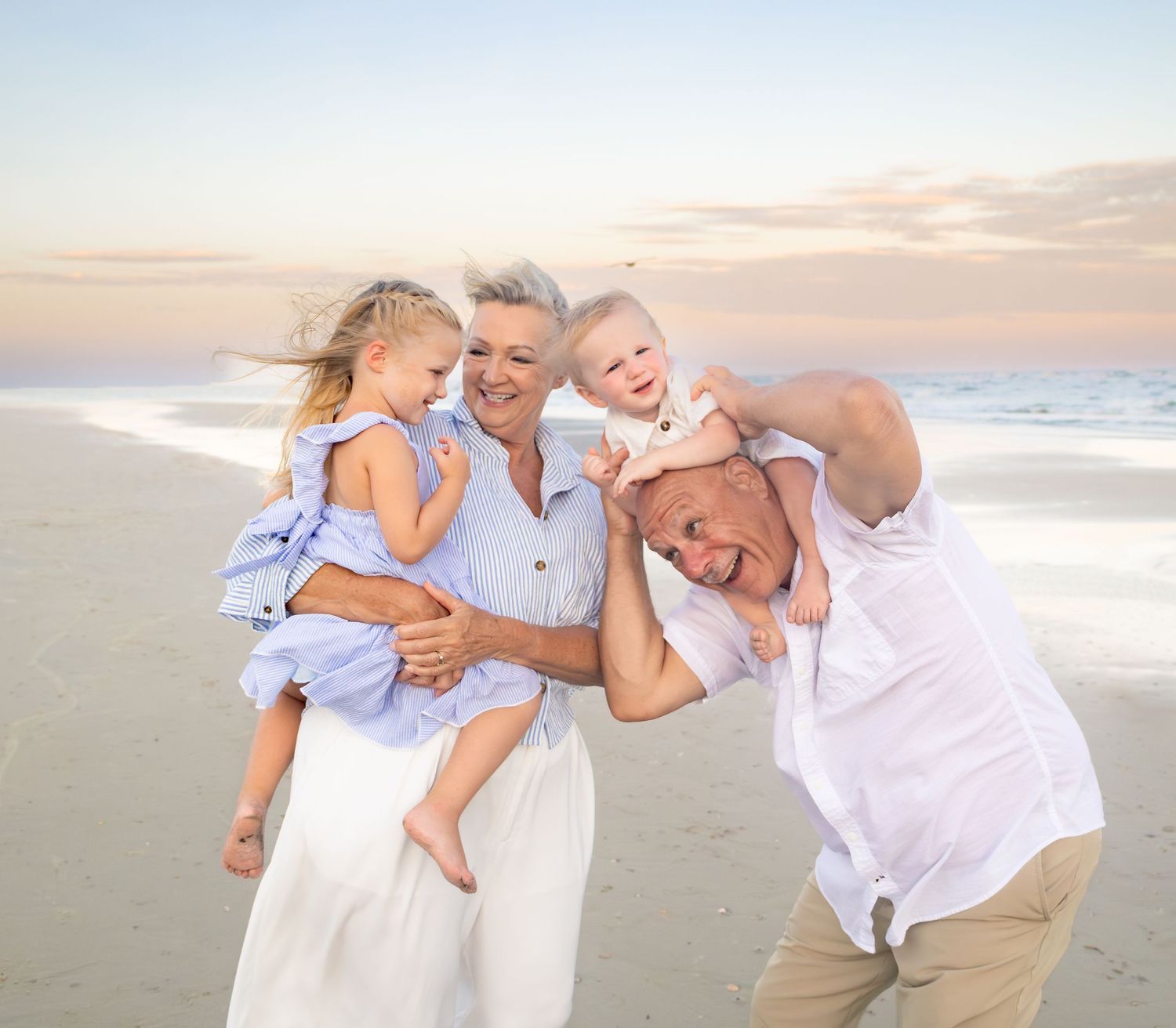 Grandparents laughing on beach at sunset holding two young grandchildren in matching blue outfits.