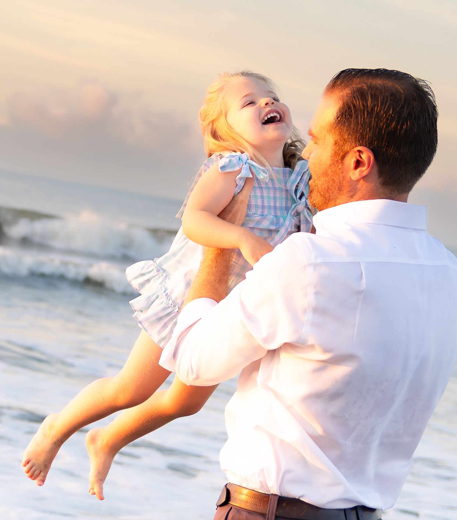 Father joyfully lifting laughing toddler girl on a sunny beach with ocean waves in the background.
