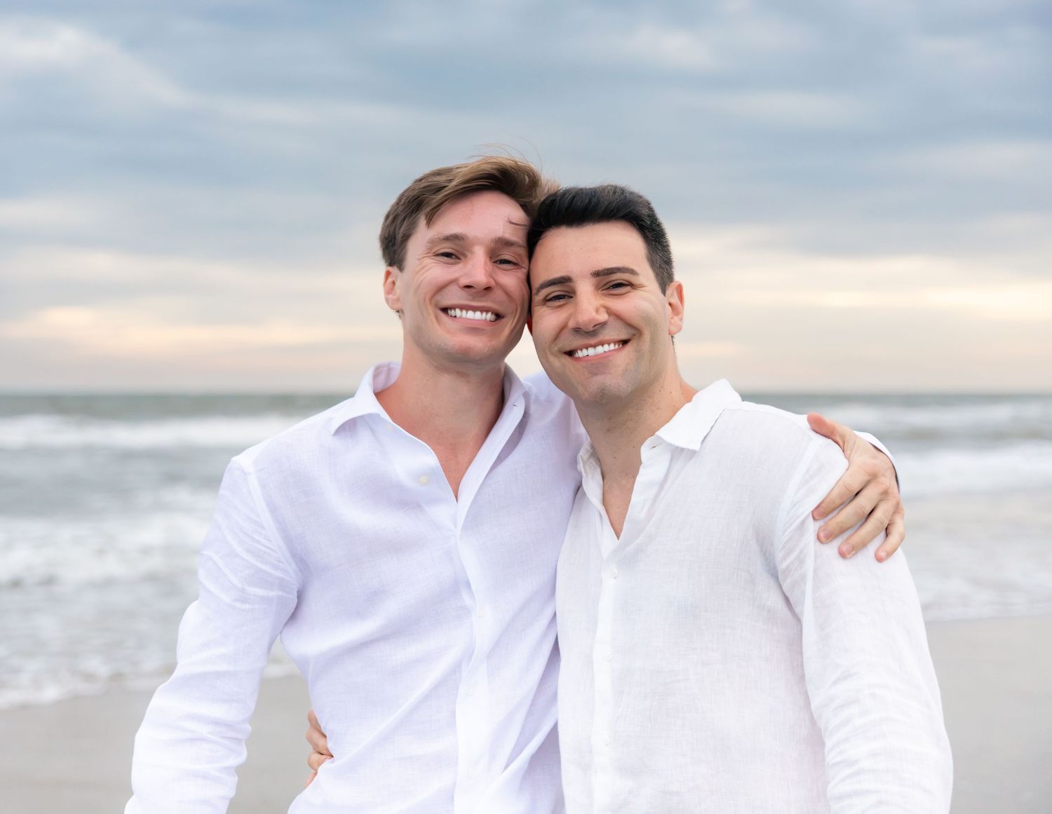 Two smiling men in white shirts embrace on a beach with ocean waves and cloudy sky behind them.