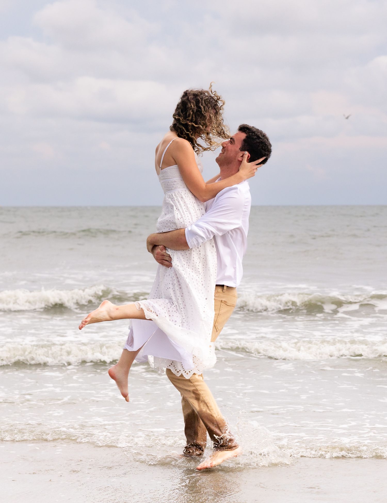 A romantic couple embraces and kisses on a beach, the man lifting the woman in a white dress by the ocean.