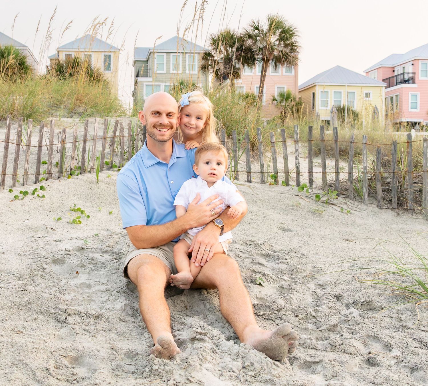 Family of three poses on sandy beach with colorful coastal homes and palm trees in the background.