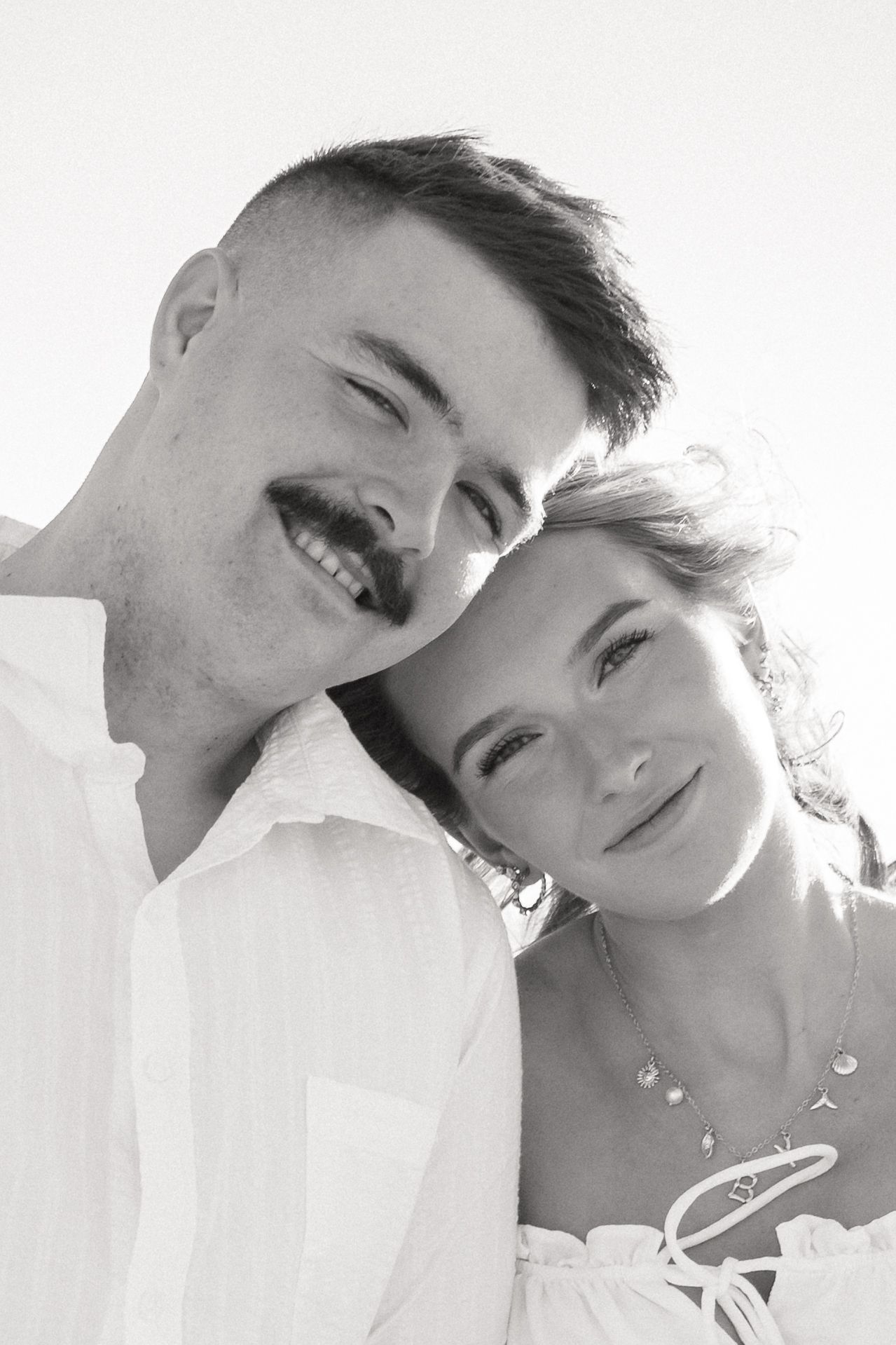 Black and white portrait of a smiling couple posing closely together outdoors in white attire.