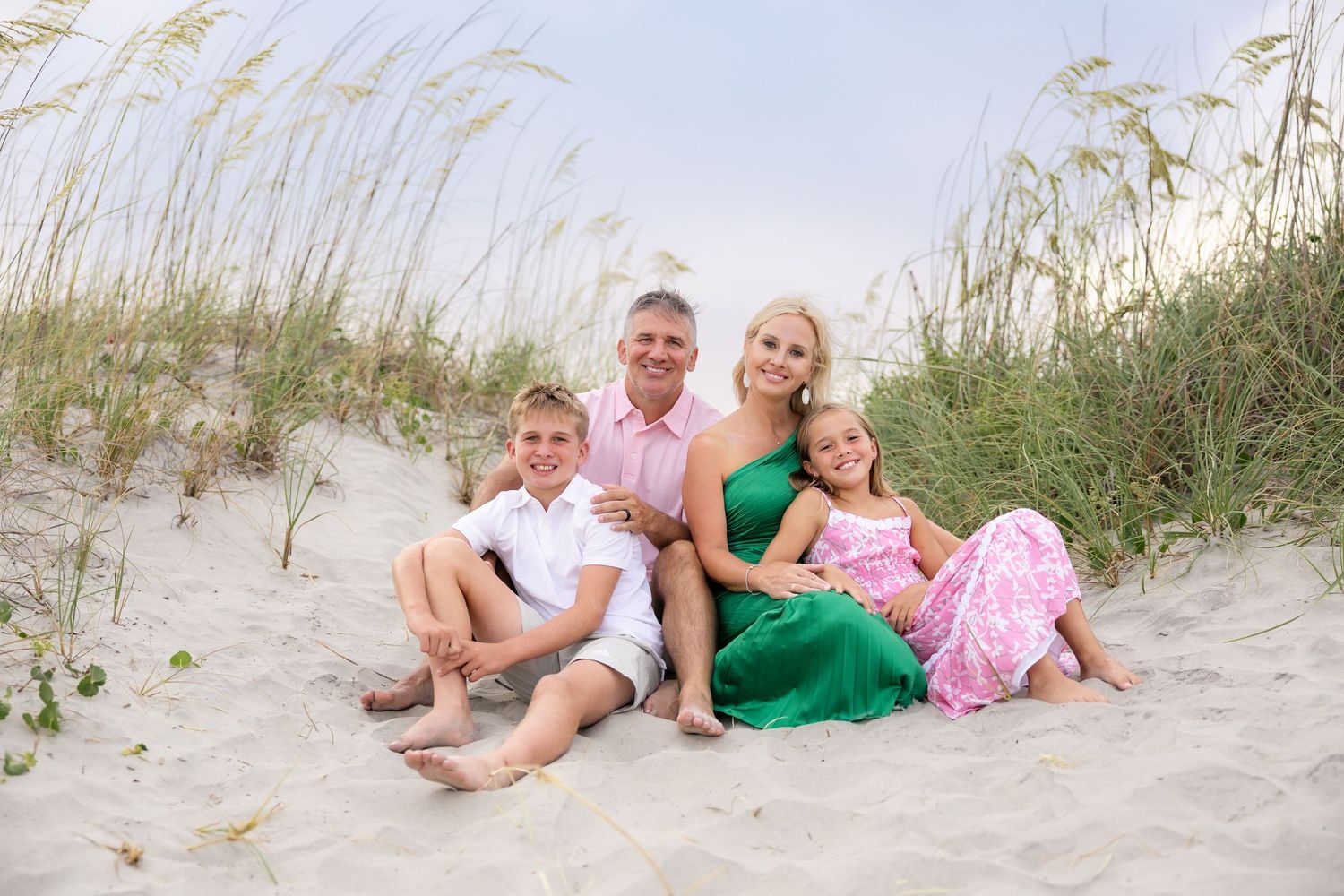 Family of four smiling together on sandy beach dunes with sea oats, wearing coordinated pink and green outfits.