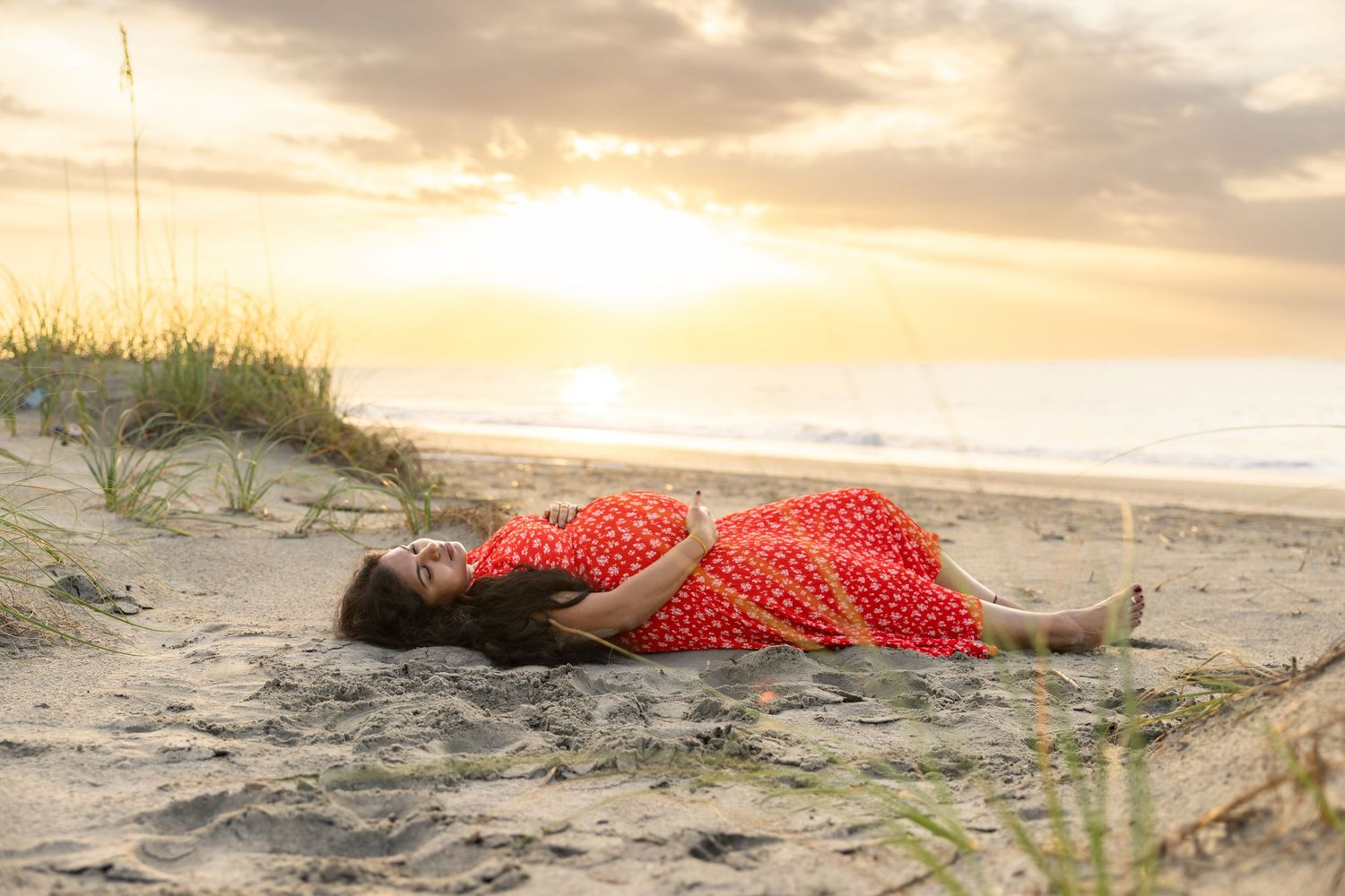 Woman in red polka dot dress lying on sandy beach dunes at golden sunset.