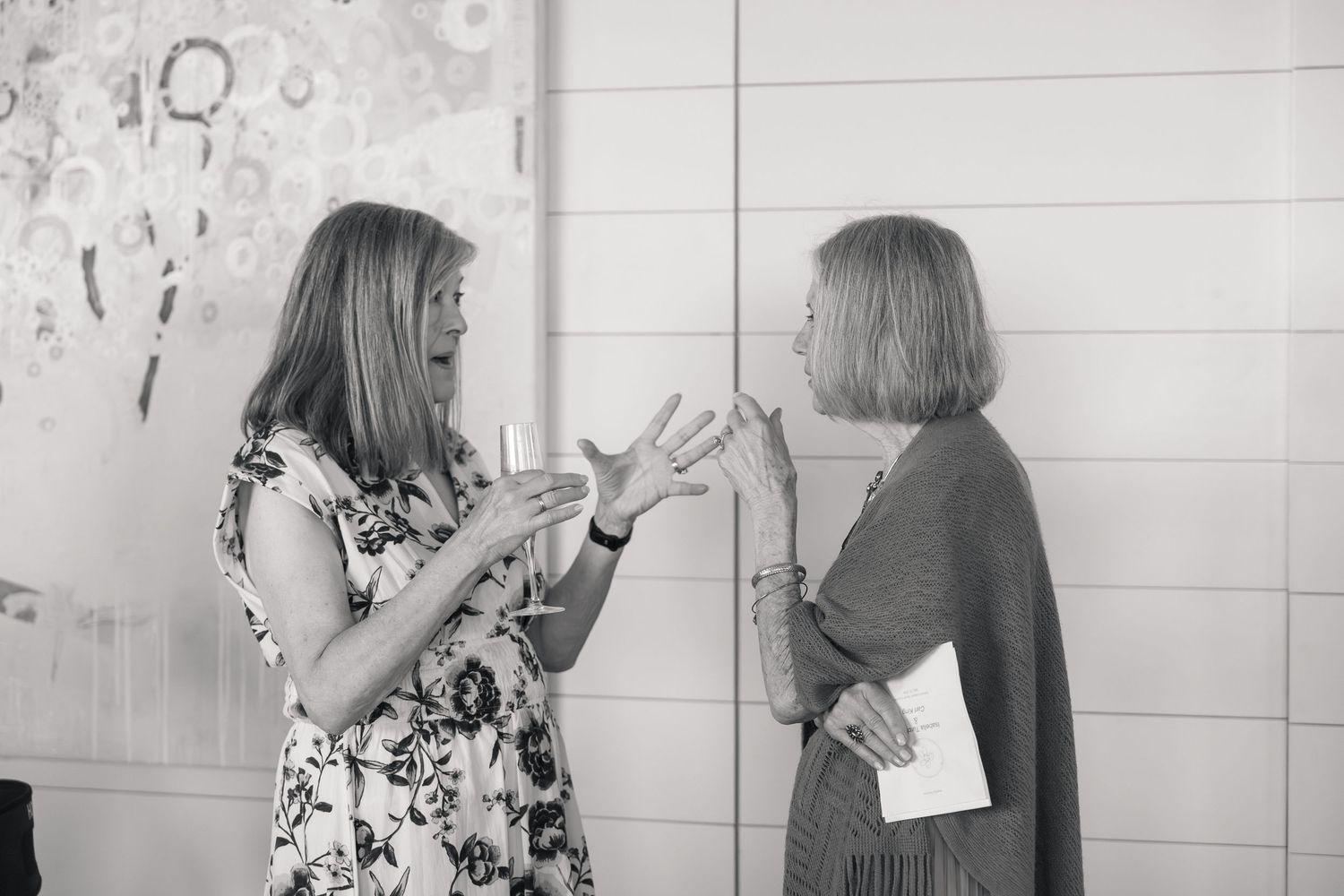 Two women engaged in conversation at an indoor event, one gesturing expressively, captured in black and white photography.