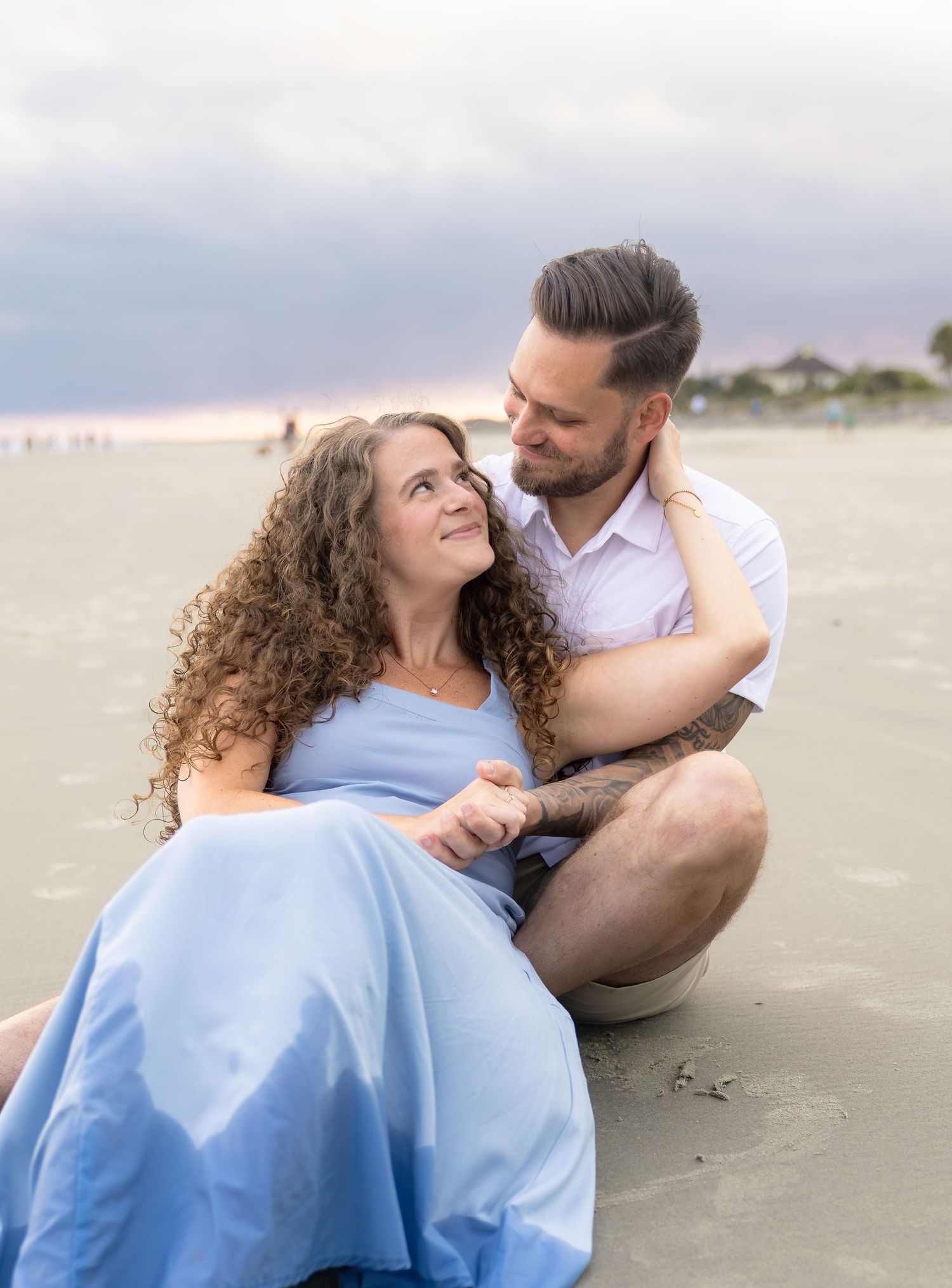 Couple sits together on beach sand, smiling at each other during golden hour engagement session.