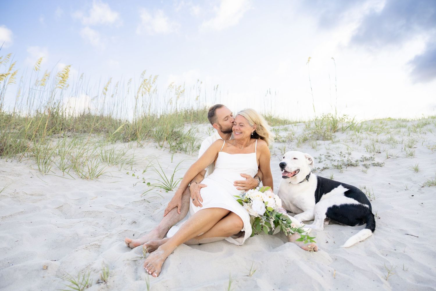 Couple in wedding attire sitting on beach sand dunes with black and white dog, holding white flowers.