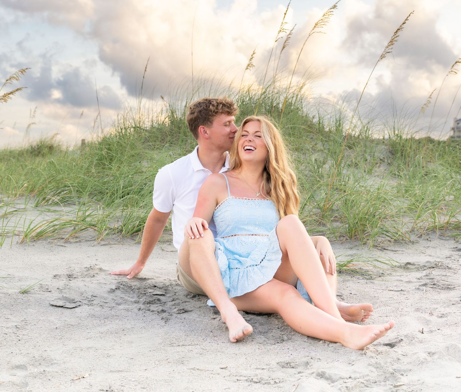 A couple sits on a sandy beach, woman in blue romper laughing as man kisses her cheek, sea oats and clouds behind.