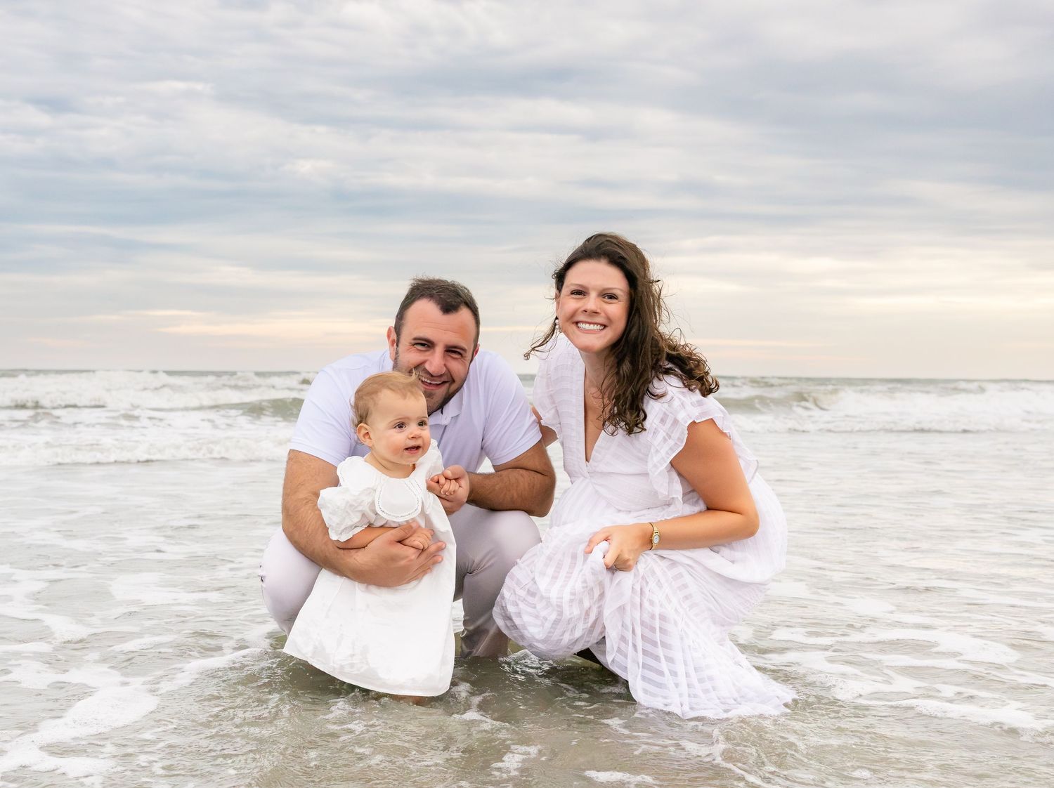 Family of three posing happily on a beach shoreline, parents in white outfits with their baby girl, ocean waves behind them.