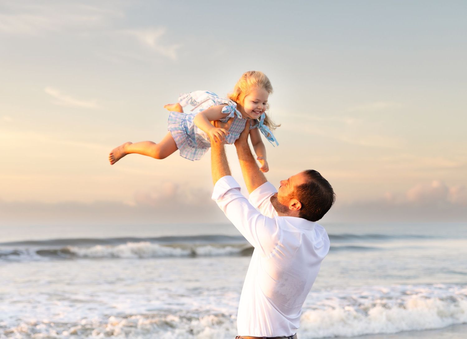 Father joyfully lifting laughing toddler into the air on a sunny beach with ocean waves in the background.